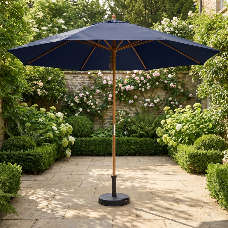 The Kratos 3m Round Wooden Parasol in Navy with UV 50+ sun protection stands on a stone terrace, surrounded by lush green plants and blooming flowers.