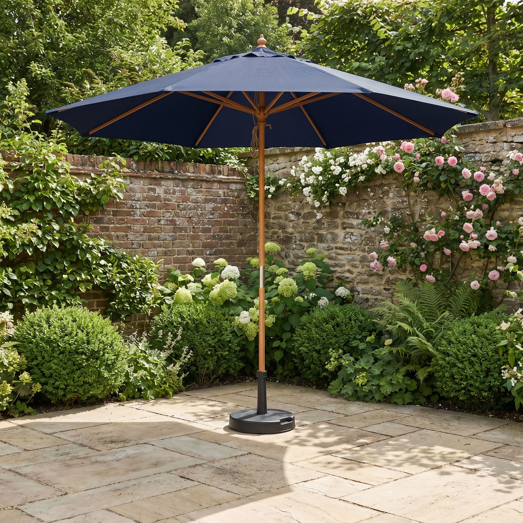 The Kratos 3m Round Wooden Parasol in Navy stands on a stone terrace, offering UV 50+ sun protection amid green bushes and blooming flowers.