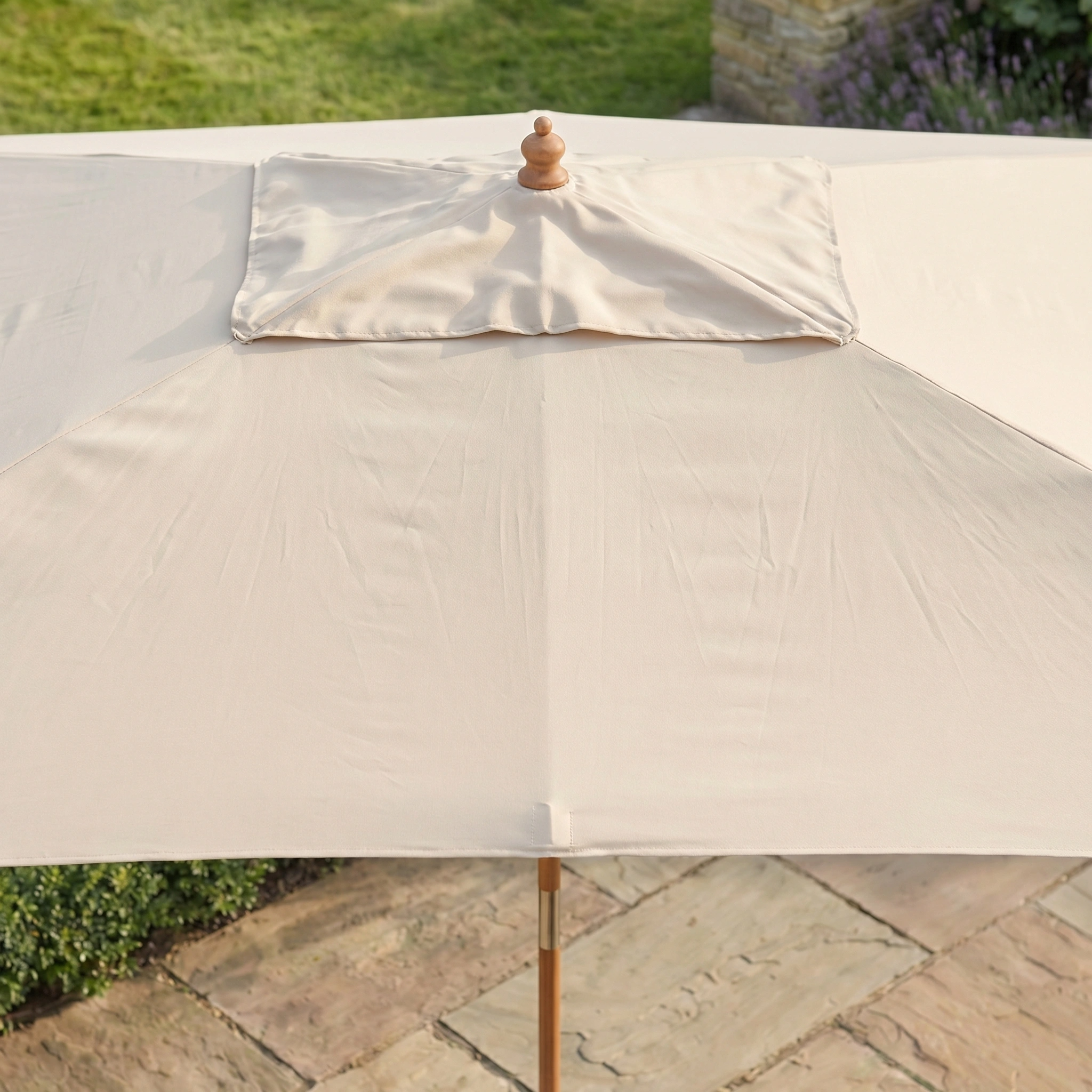 The Kratos 3m x 2m Rectangular Wooden Parasol in Beige offers stylish UV 50+ sun protection, featuring a cream finish and sturdy wooden frame, perfect for placement on stone tiles amid grass and plants.