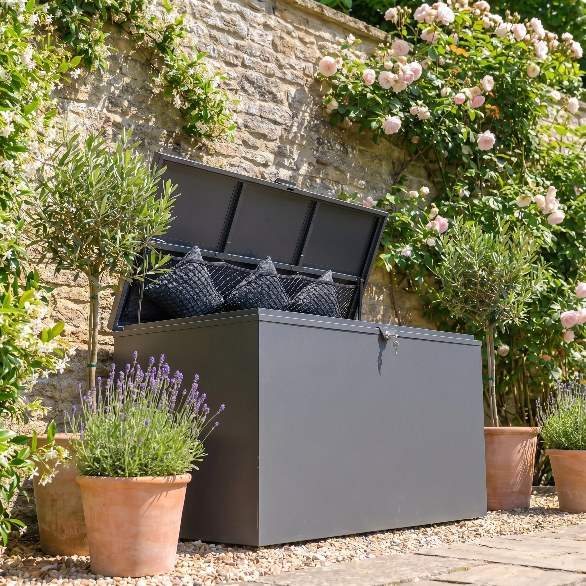 The Large Storage Box in Grey, made from galvanized steel with an open lid, holds cushions and sits among potted plants and blooming flowers—ideal for stylish outdoor storage.