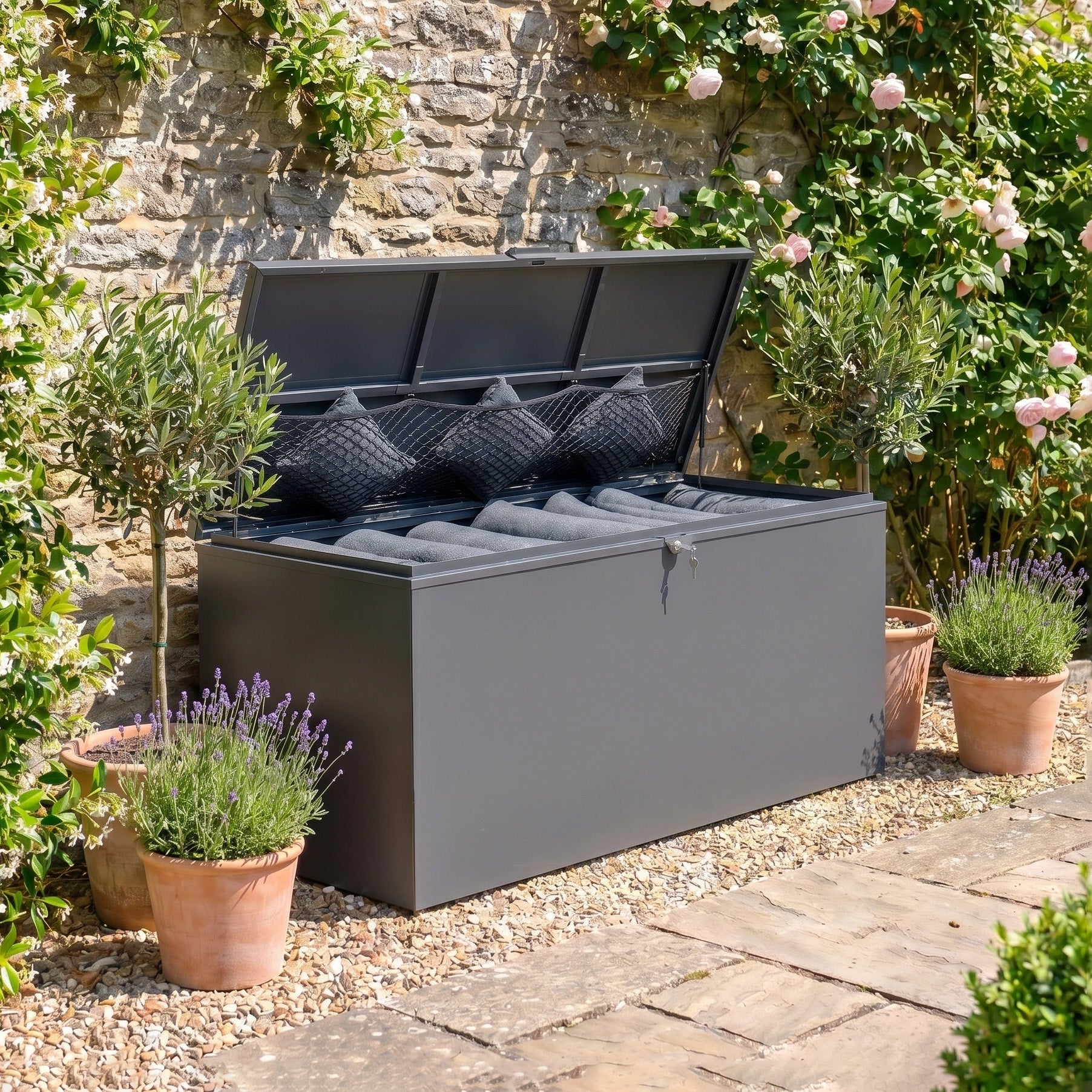 The Large Storage Box in Grey is a spacious outdoor bench with black cushions and a galvanised steel frame, perfect for stylish storage on your patio among potted plants and flowers.