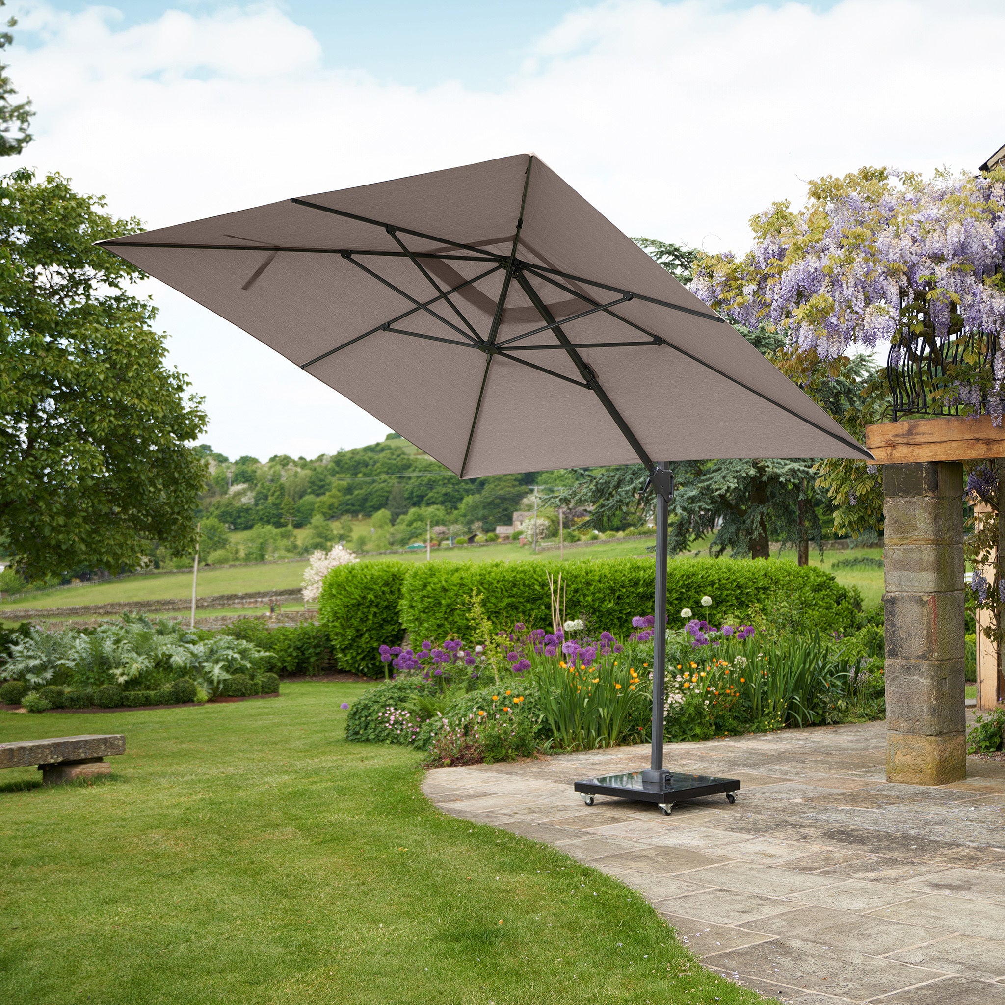The Pacific Lifestyle Challenger T2 3m Square Cantilever Parasol in Taupe, featuring a powder-coated aluminum frame, enhances the patio's elegance while complementing the garden and background trees.
