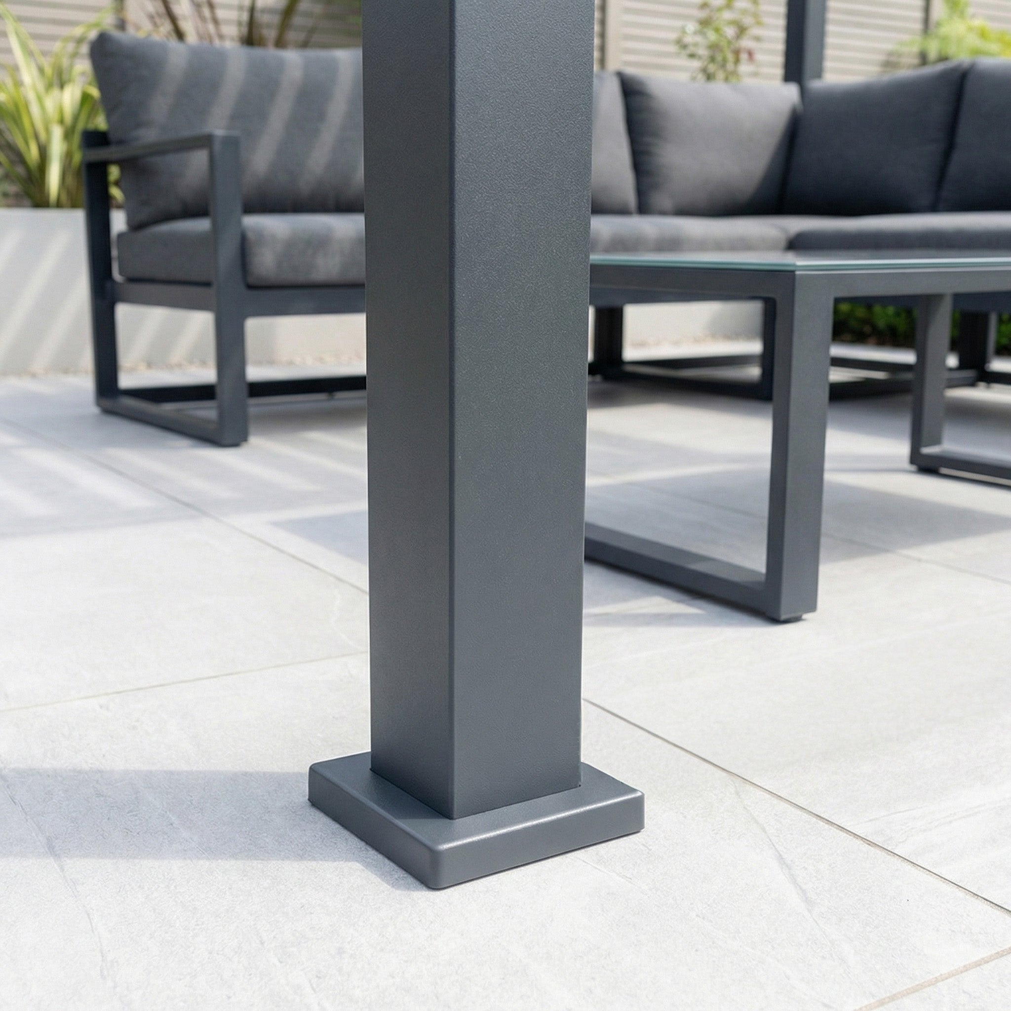 Close-up of a metal post on a tiled patio, part of the PergoSTET Lite Neo 3m x 7m Metal Rectangular Pergola in Grey with adjustable louvres, outdoor furniture, and plants visible in the background.