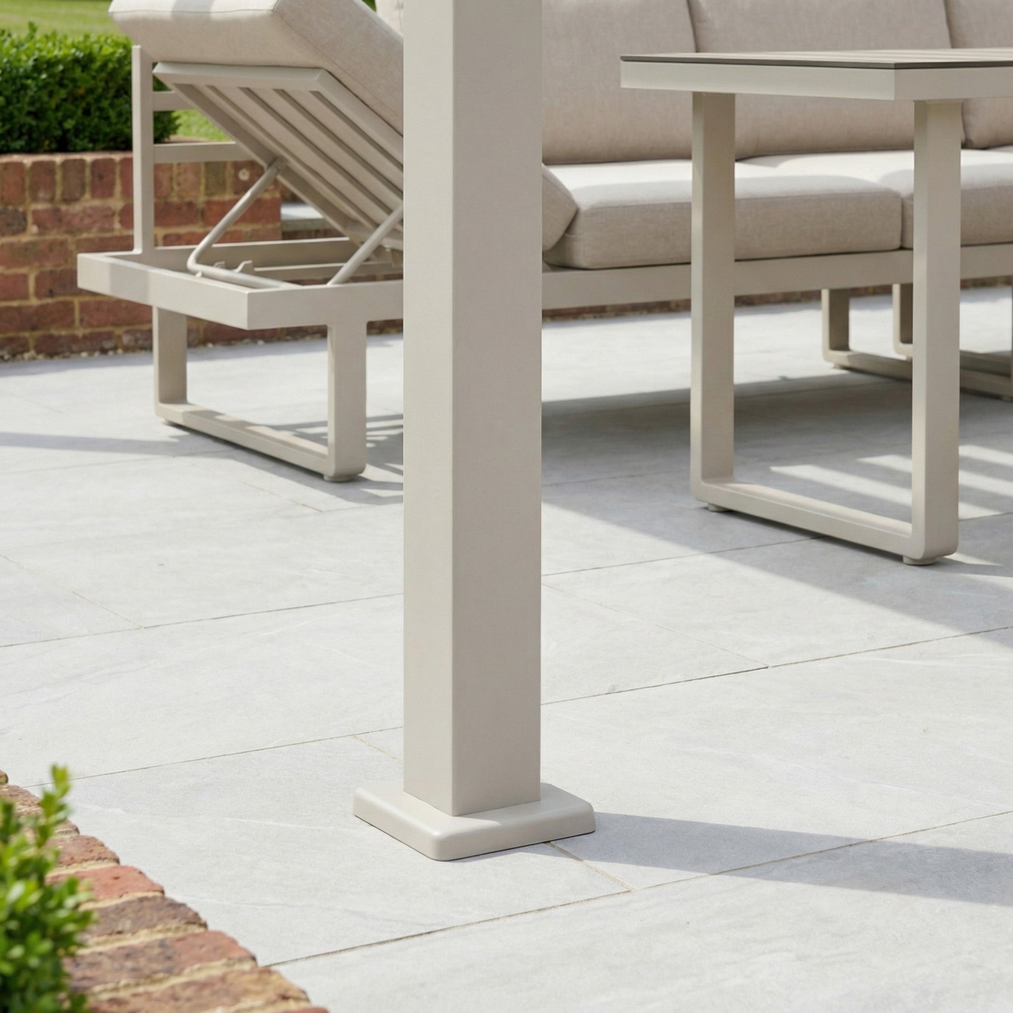 Outdoor patio furniture with beige metal frames sits on gray tile flooring, beside grass and a brick border, beneath the PergoSTET Lite 3m x 4m Metal Rectangular Pergola in Latte, which features tilting louvres for adjustable shade.