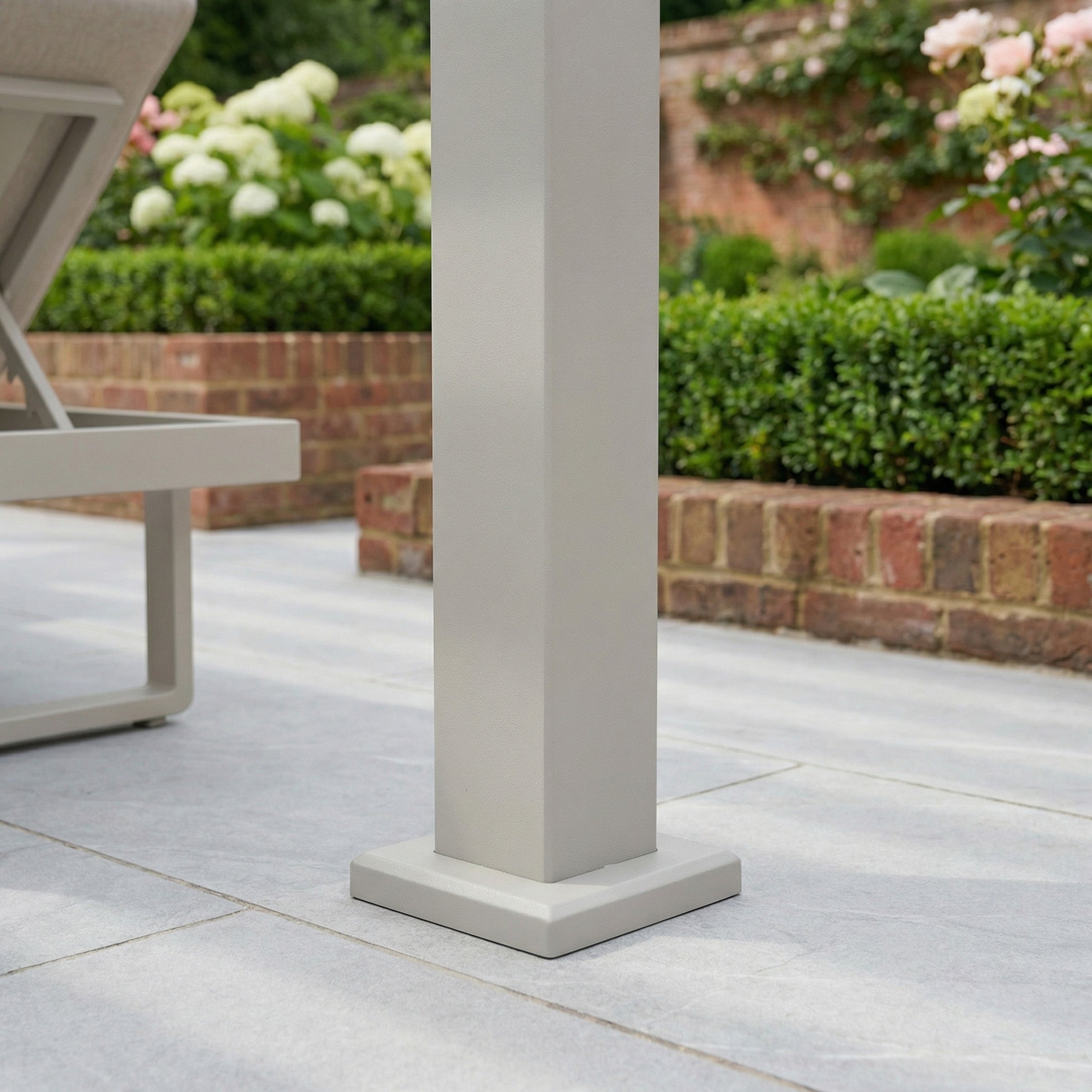 The PergoSTET Lite 3m x 4m Metal Rectangular Pergola in Latte stands on a tiled patio, surrounded by lush garden plants and a lounge chair—ideal for outdoor shade with optional tilting louvres.