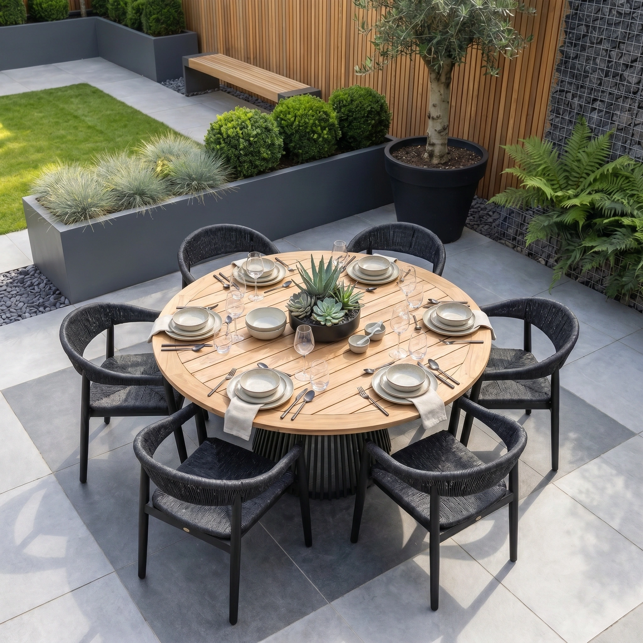 The Lucca 6 Seat Dining Set with Teak Round Table in Black is ideal for modern outdoor dining, pairing well with lush garden plants and wooden privacy fencing.