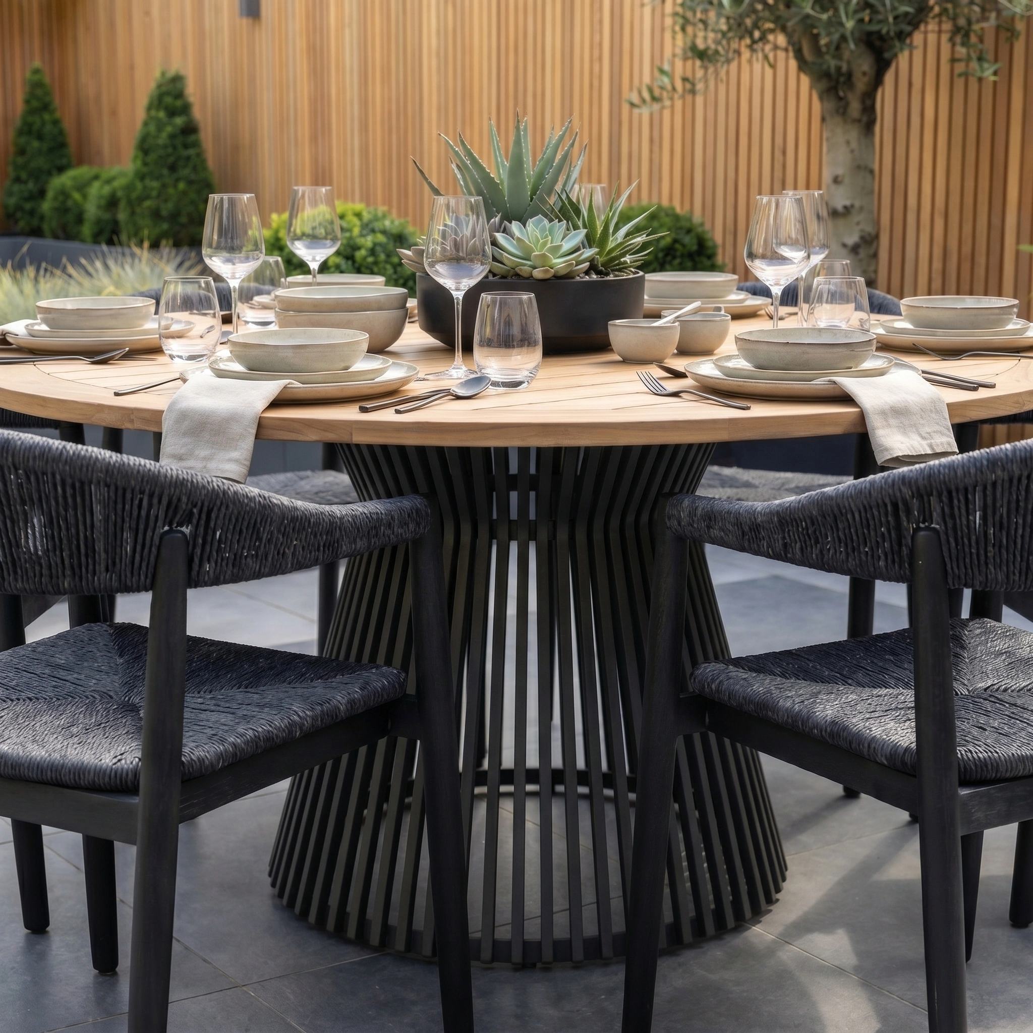The Lucca 6 Seat Dining Set with Teak Round Table in Black is styled with dishes, glasses, napkins, and a succulent centerpiece—ideal outdoor dining furniture for gatherings.
