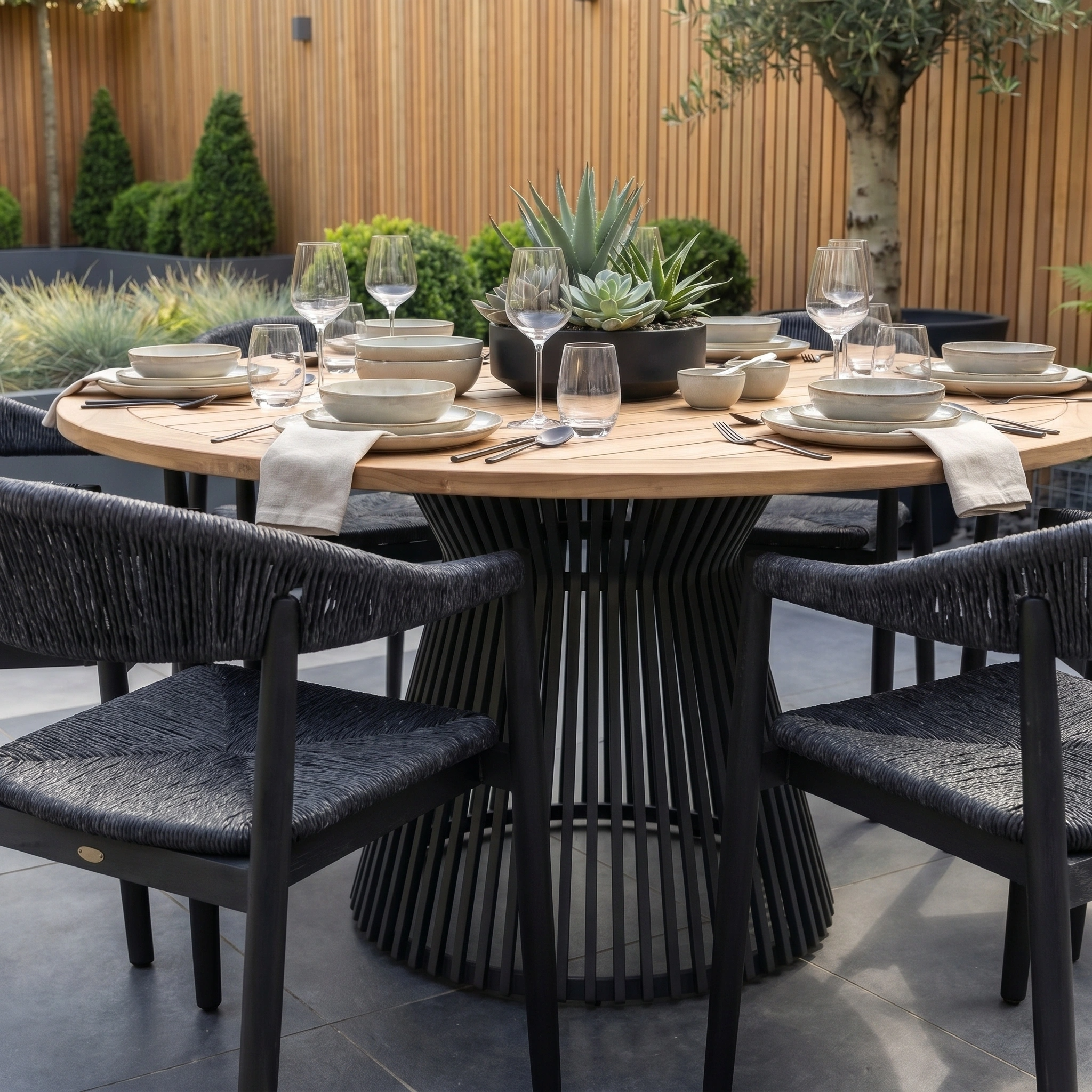The Lucca 6 Seat Dining Set with Teak Round Table in Black is arranged for six, styled with neutral tableware and a succulent centerpiece, creating an inviting outdoor dining setup on the patio.