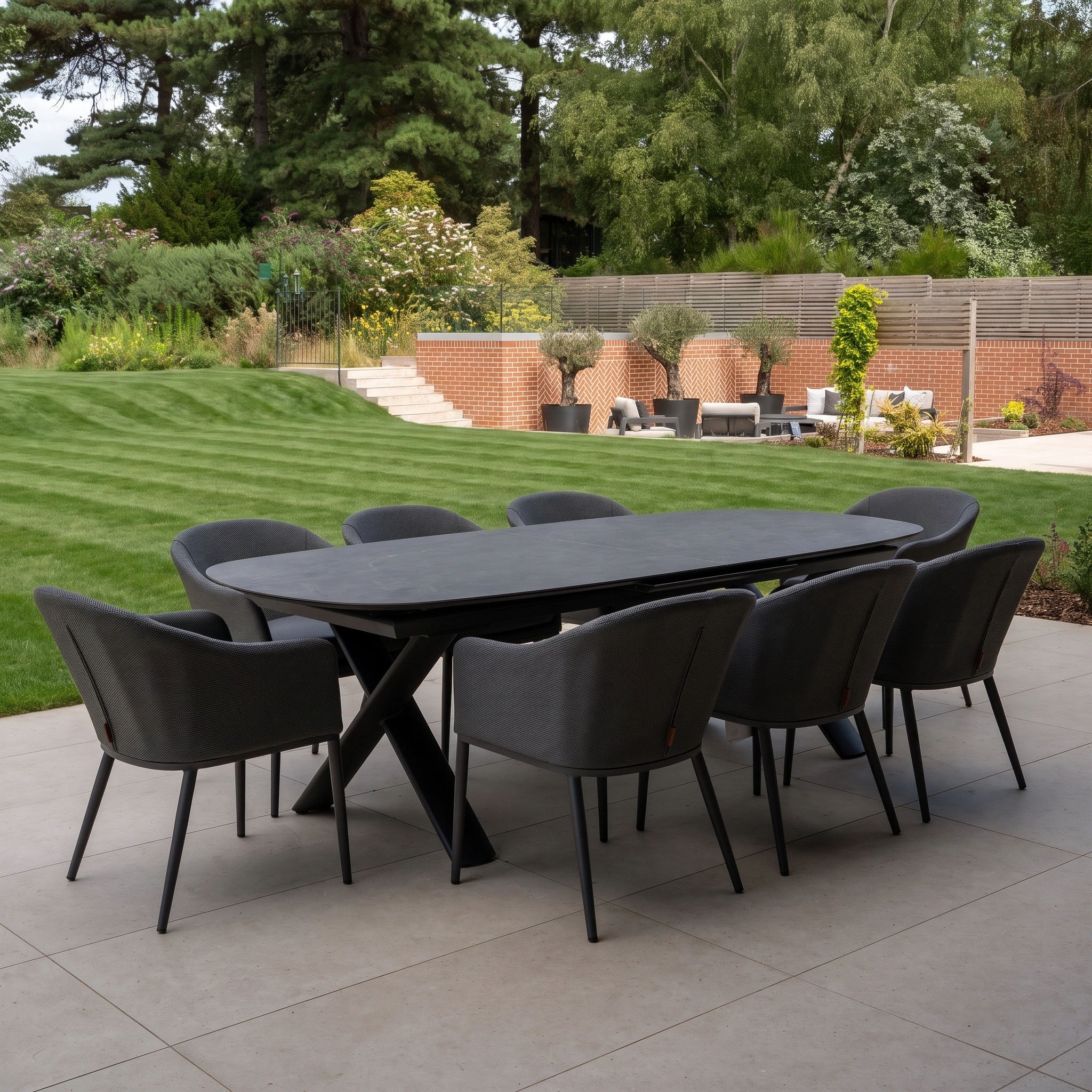 The Luna 8 Seat Outdoor Fabric Oval Dining Set with Albatross Extending Table in Grey features eight chairs arranged on a patio, overlooking a green lawn and garden.