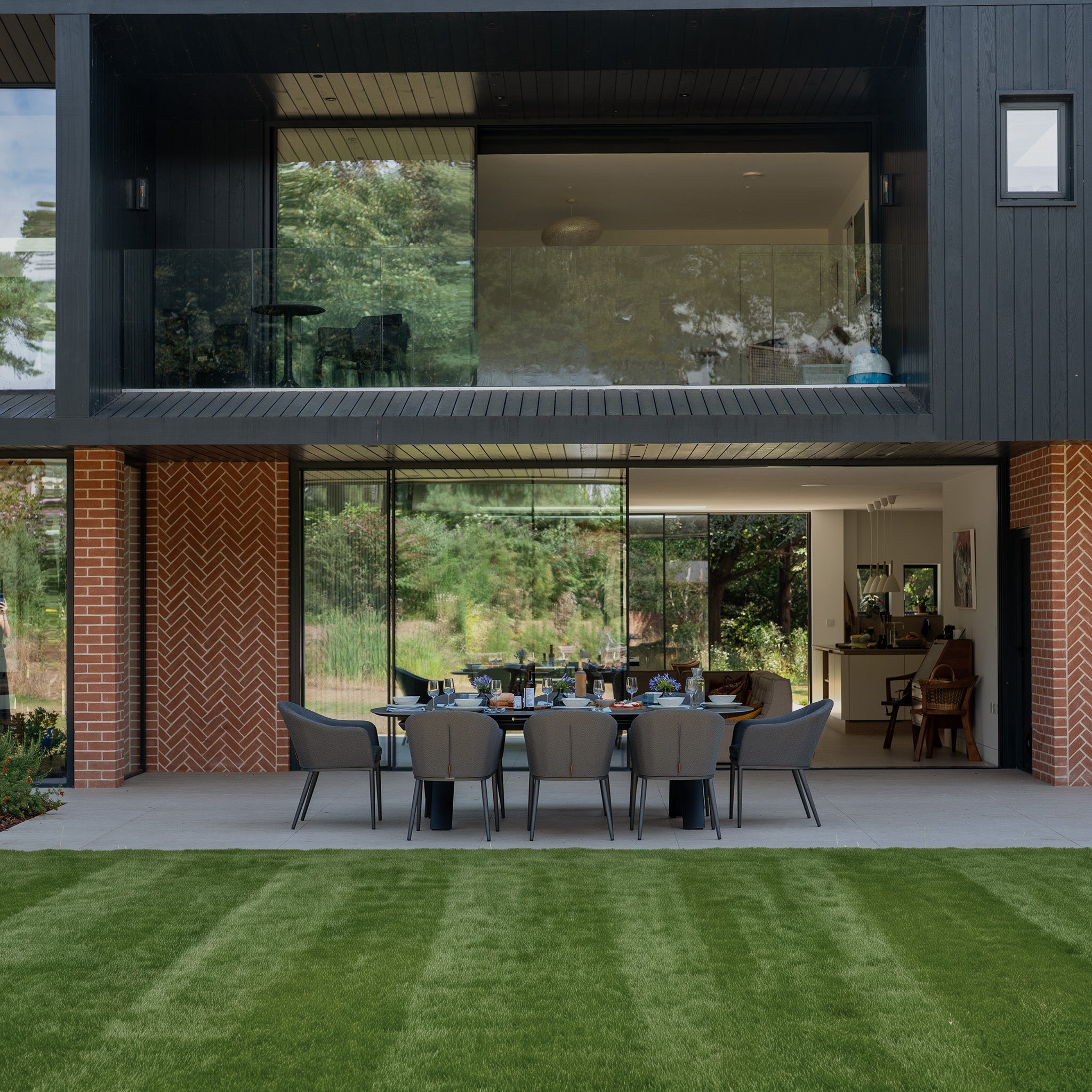 A modern house with large glass windows features the Luna 8 Seat Outdoor Fabric Oval Dining Set with Albatross Extending Table in Grey, crafted from weather-resistant garden furniture, set on a lush green lawn in the foreground.
