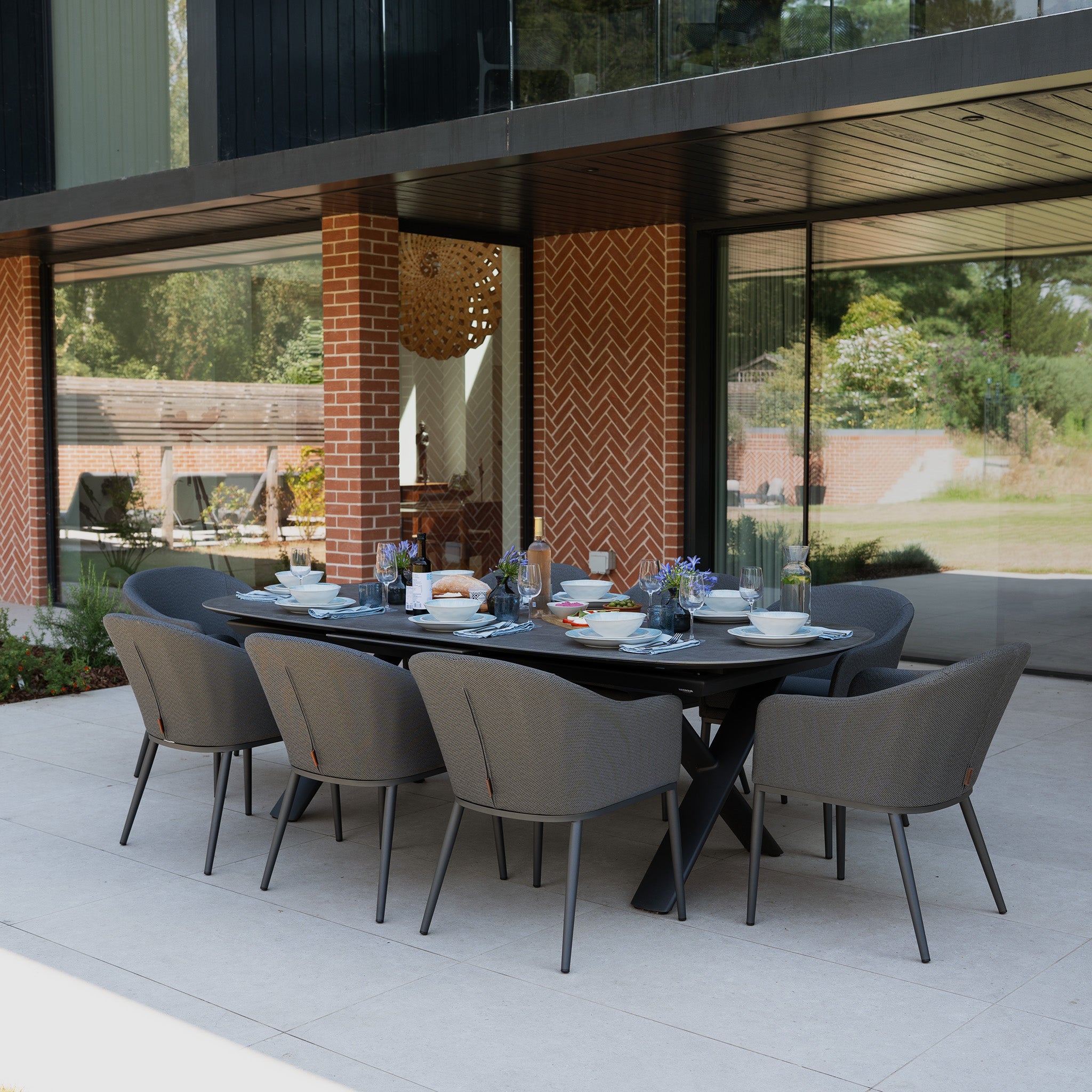 The Luna 8 Seat Outdoor Fabric Oval Dining Set with Albatross Extending Table in Grey is arranged for a meal near a modern house with large windows, featuring eight weather-resistant gray chairs.