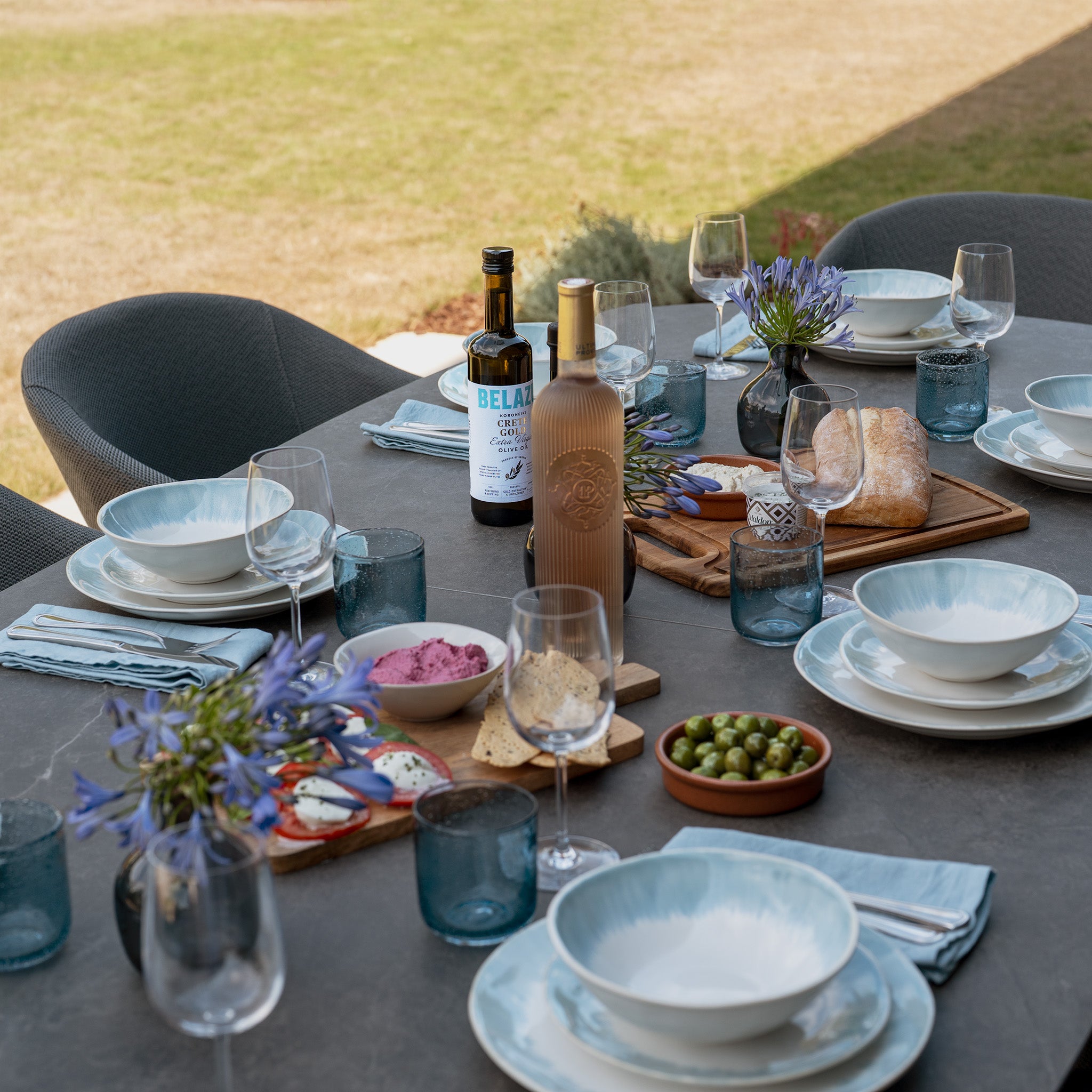 The Luna 8 Seat Outdoor Fabric Oval Dining Set with Albatross Extending Table in Grey offers weather-resistant garden furniture that combines durability and style, shown here arranged with plates, drinks, bread, olives, and flowers on the spacious extending table—perfect for any outdoor meal.