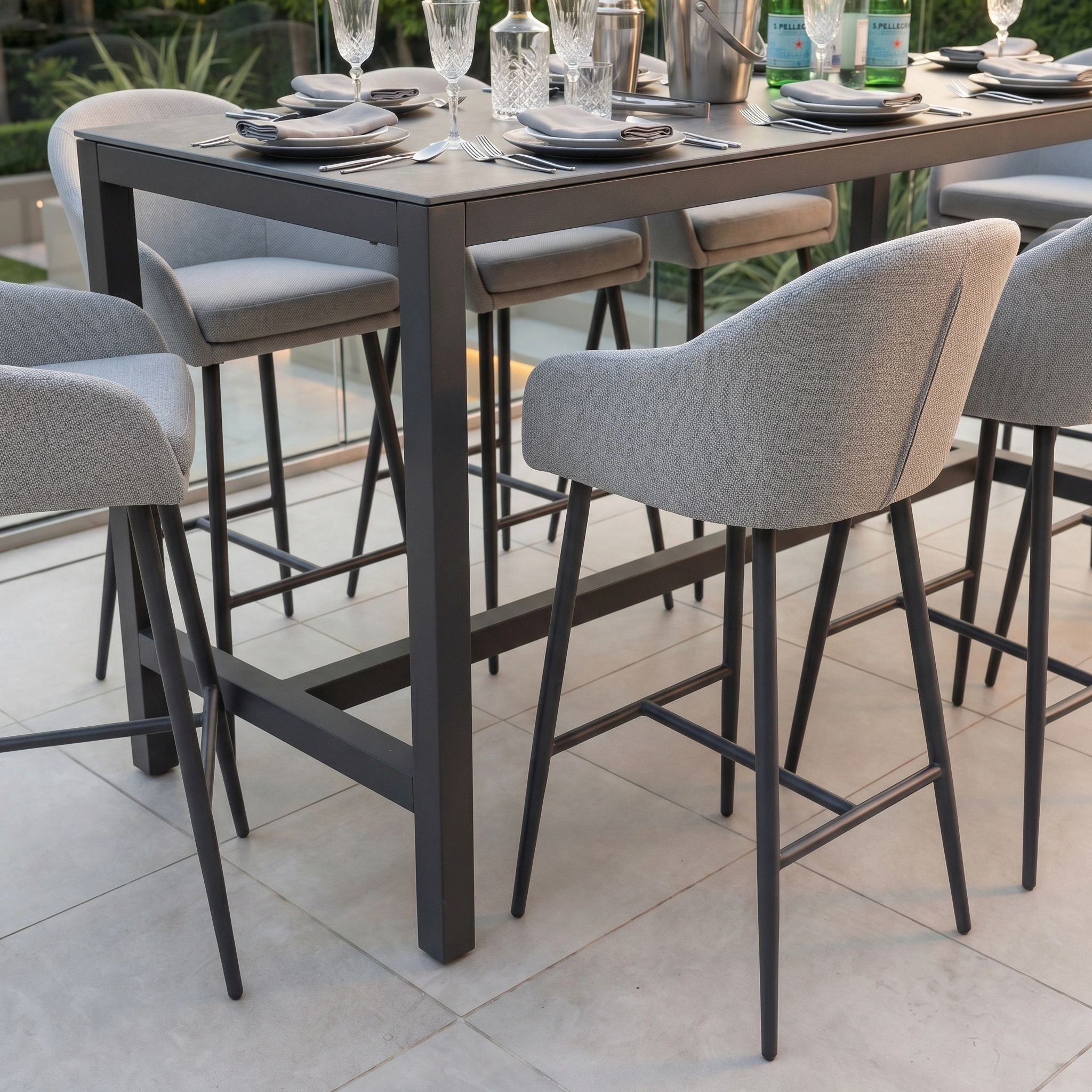 The Luna 8 Seat Bar Set in Oyster Grey features a modern outdoor dining table and eight gray cushioned chairs made from all-weather material. Complete with plates, glasses, and cutlery, it ensures durability and comfort for any outdoor gathering.