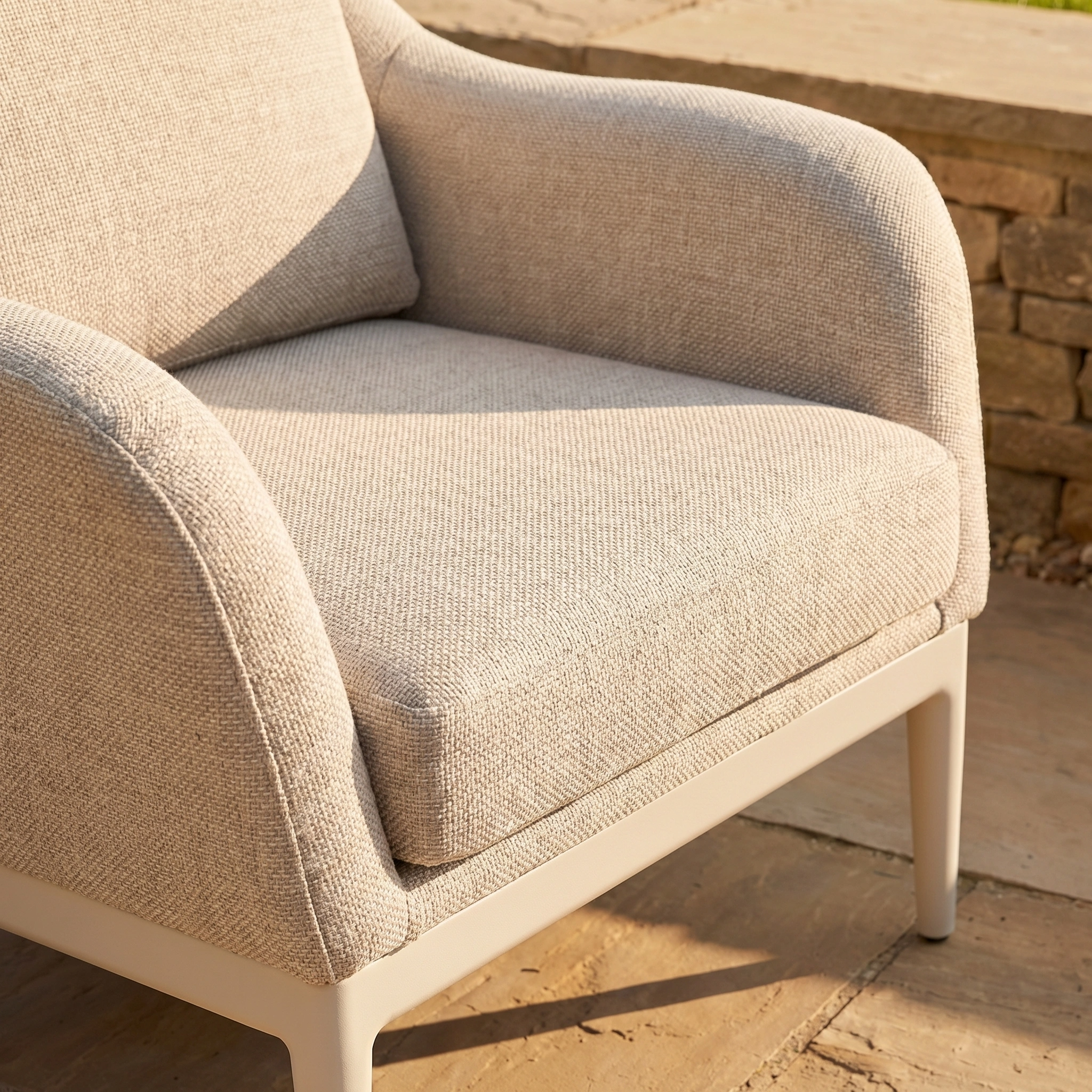 The Luna Armchair in Fawn, upholstered in stain-proof outdoor fabric, sits on a stone patio with sunlight casting soft shadows across its seat and armrest.