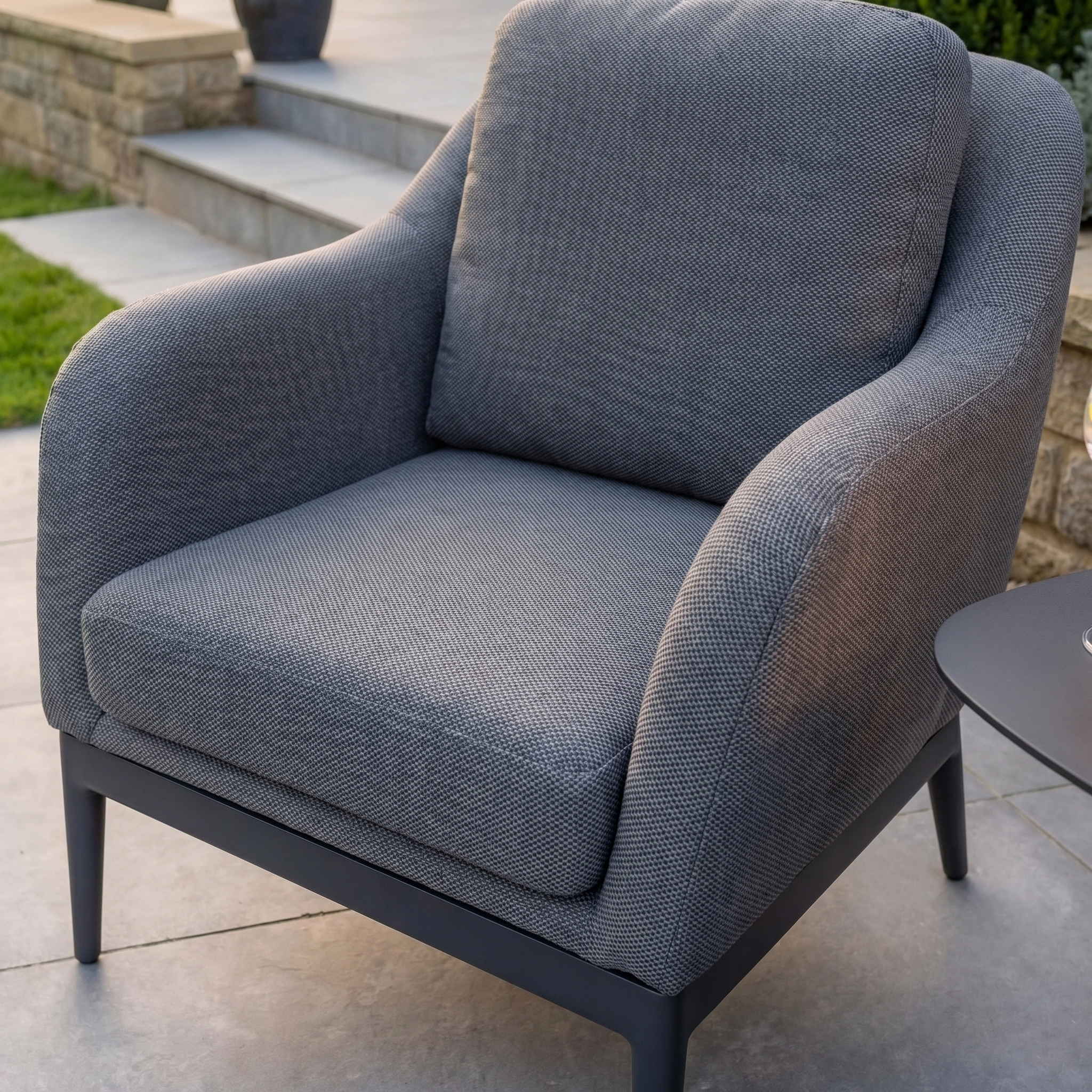 The Luna Armchair in Grey, crafted for all-weather durability and outdoor comfort, sits on a patio near stone steps and a garden.