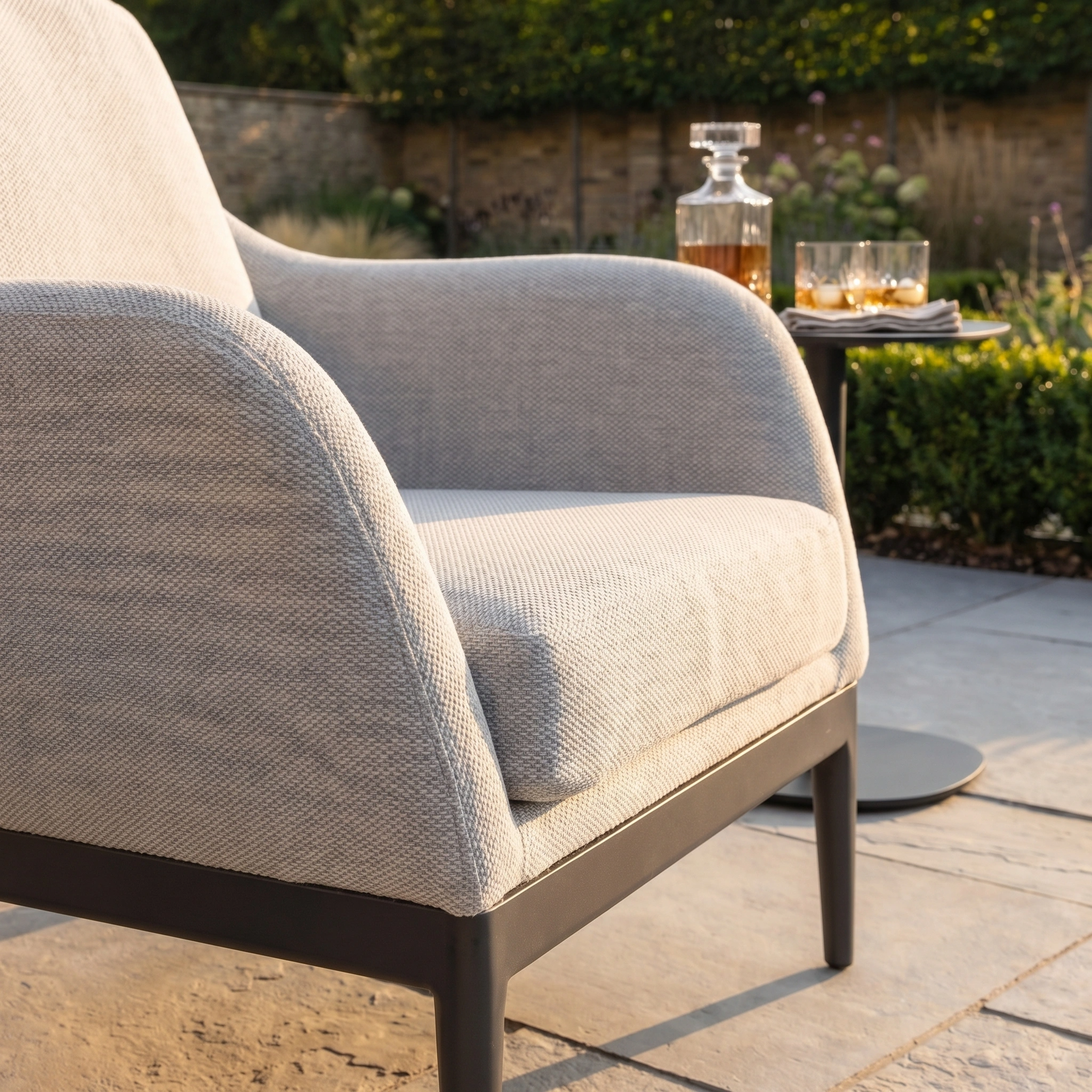 The Luna Armchair in Oyster Grey, made from all-weather material, is placed on a patio next to a small table with a decanter and glasses, basking in sunlight.
