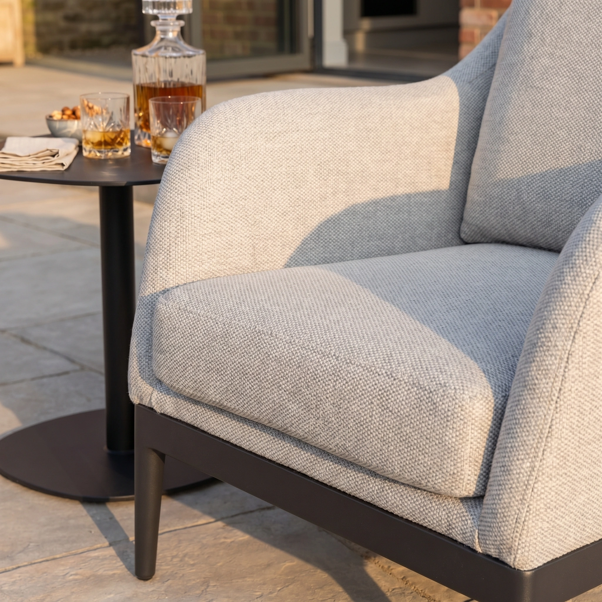 The Luna Armchair in Oyster Grey, made with all-weather material, sits outdoors next to a small table holding whiskey glasses and a decanter.