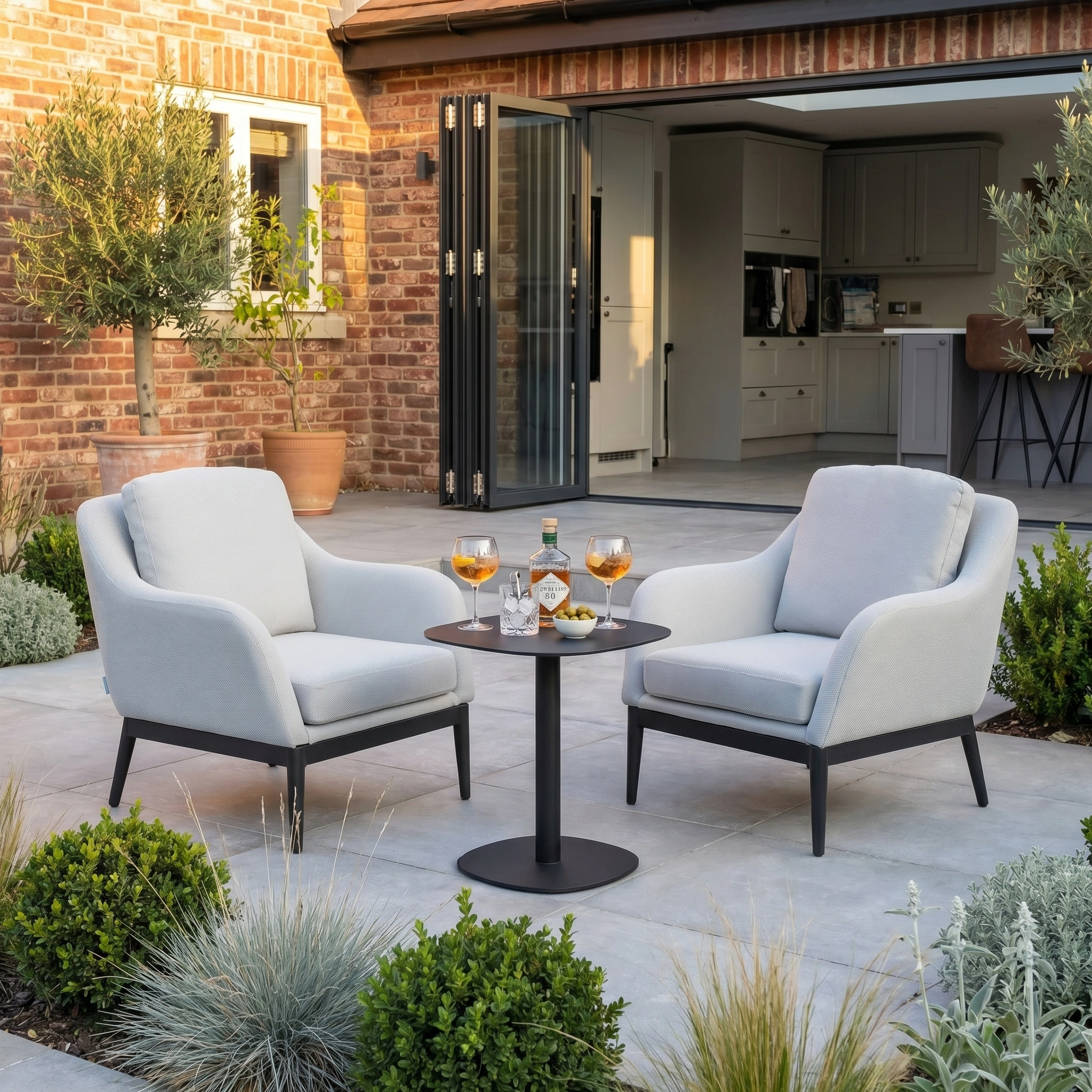 Two Luna Armchairs in Oyster Grey with all-weather cushions and a small table with drinks sit in a modern garden outside a brick house.