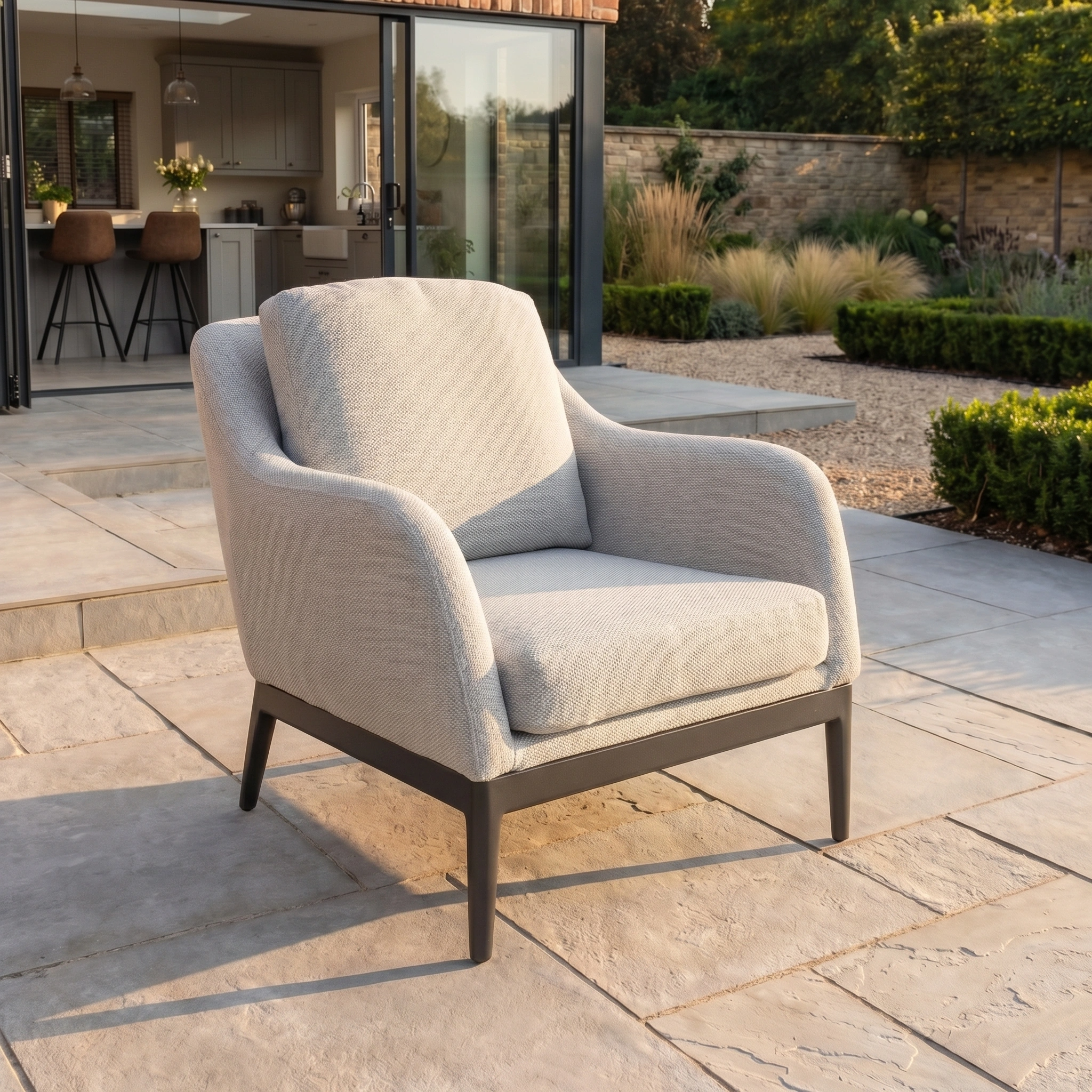 The Luna Armchair in Oyster Grey, made with all-weather material, is placed on a stone patio outside a modern home featuring an open kitchen in the background.
