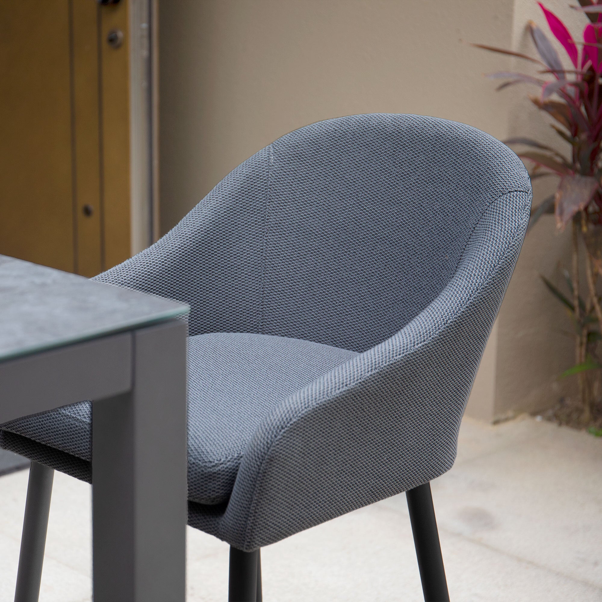 The Luna Outdoor Fabric Bar Stool in Grey (Ex Display) with black legs is placed outdoors by a table near a beige wall and plants.