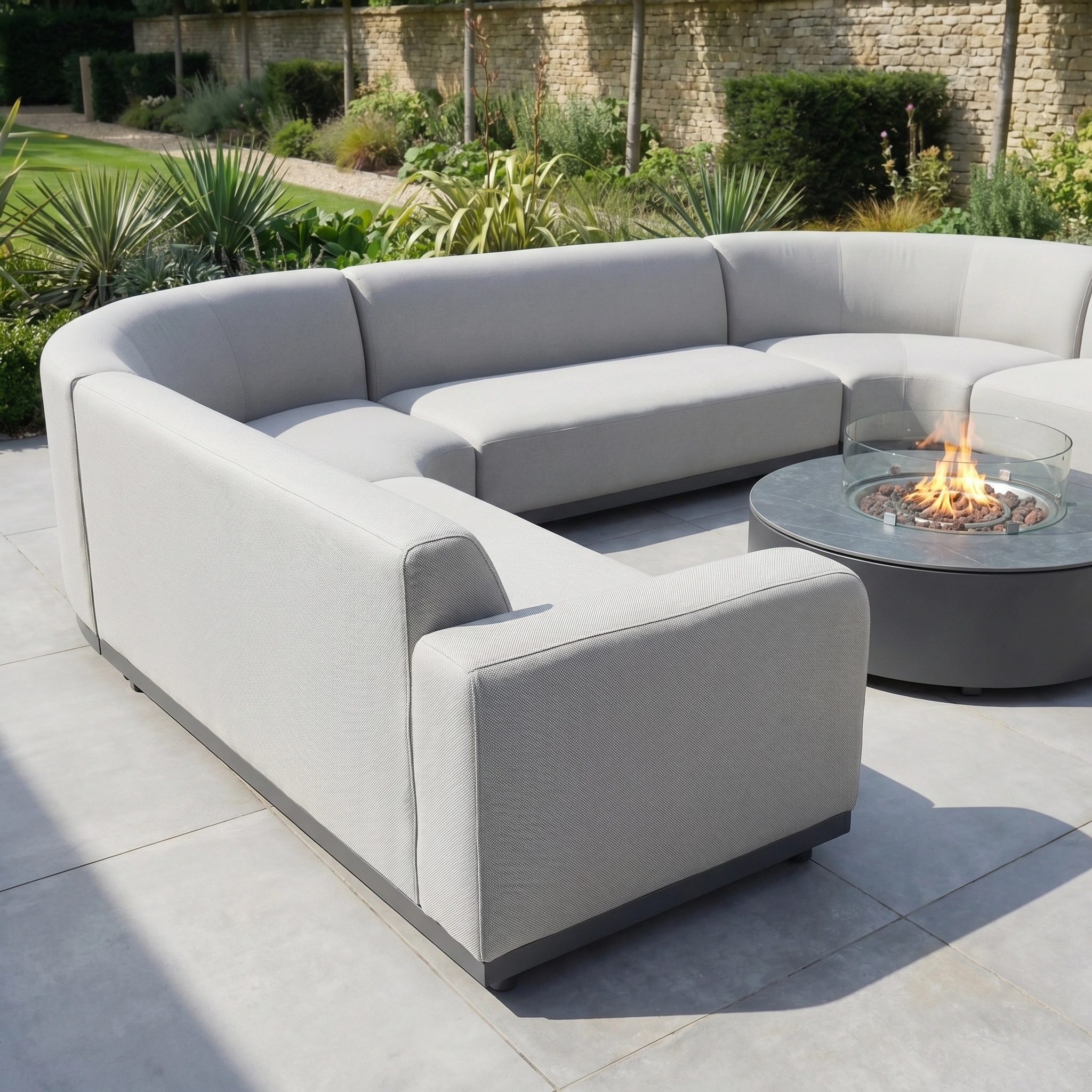The Luna U-Shape Outdoor Fabric Curved Corner Group with Round Firepit Coffee Table in Oyster Grey sits on a stone patio, enhanced by a garden and stone wall in the background.