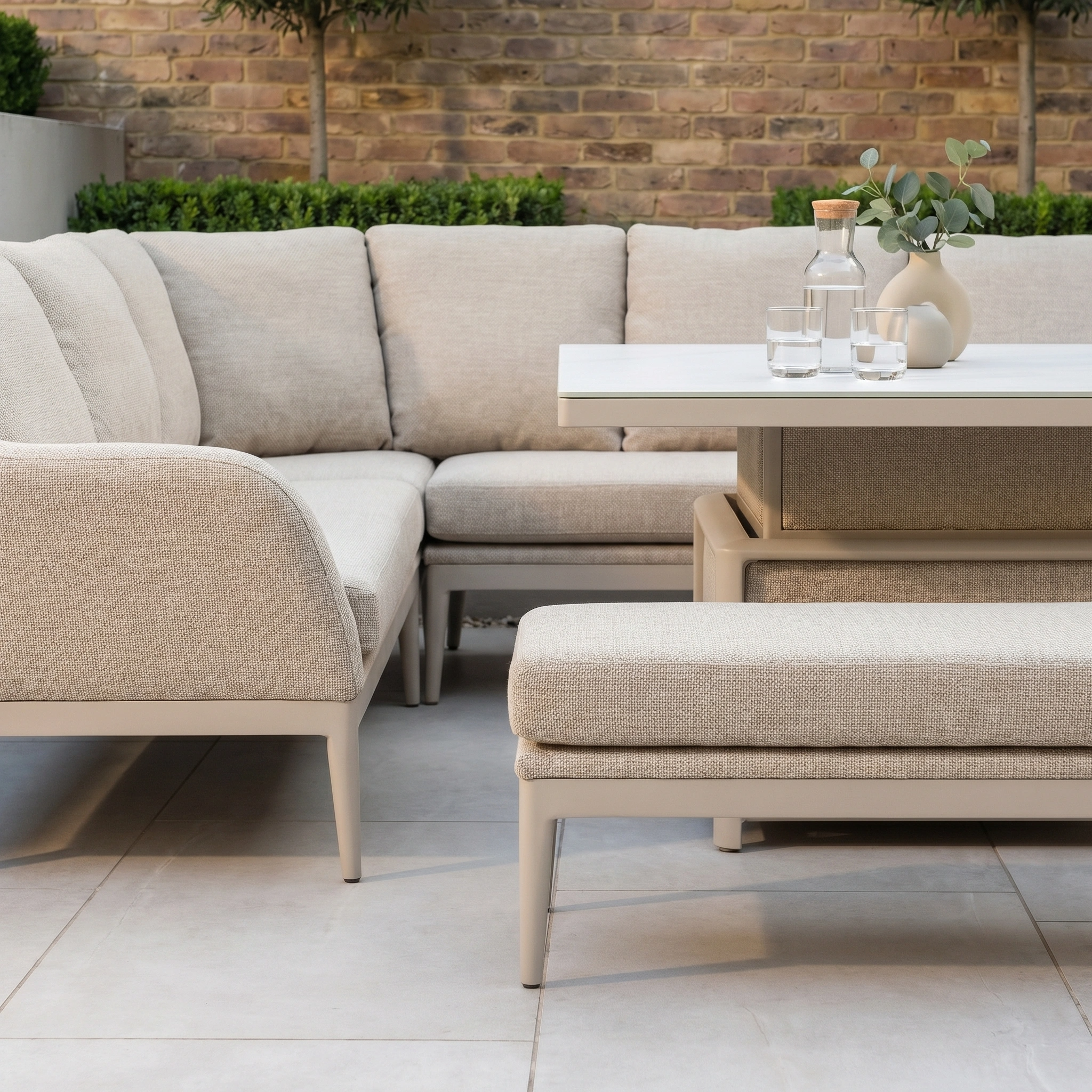 A modern patio features the Luna Deluxe Outdoor Fabric Square Corner Dining Set with Rising Table in Fawn, complemented by glassware and potted plants against a brick wall.
