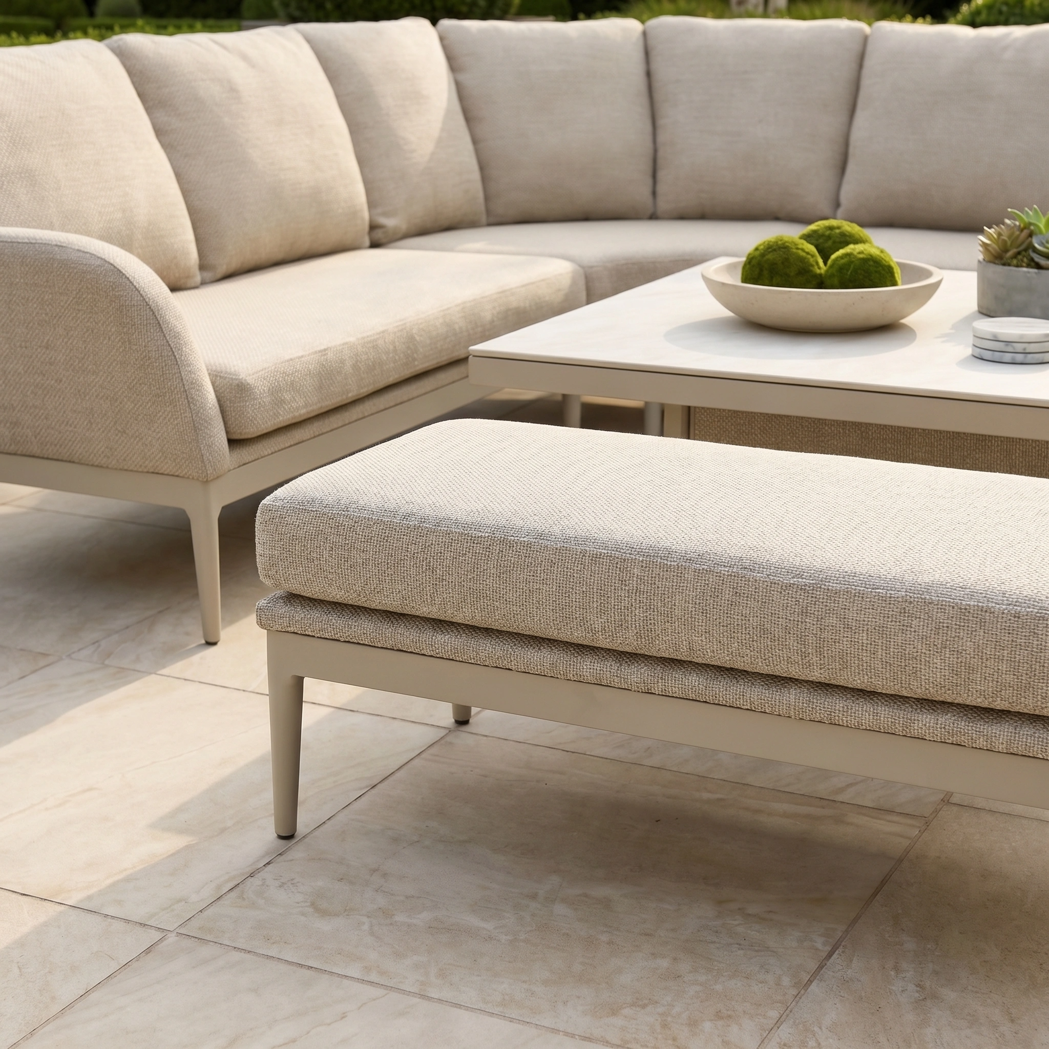 The Luna Deluxe Outdoor Fabric Square Corner Dining Set in Fawn features a sectional sofa and ottoman on a tiled patio, complemented by a rising table with a bowl of moss balls for an inviting outdoor dining experience.