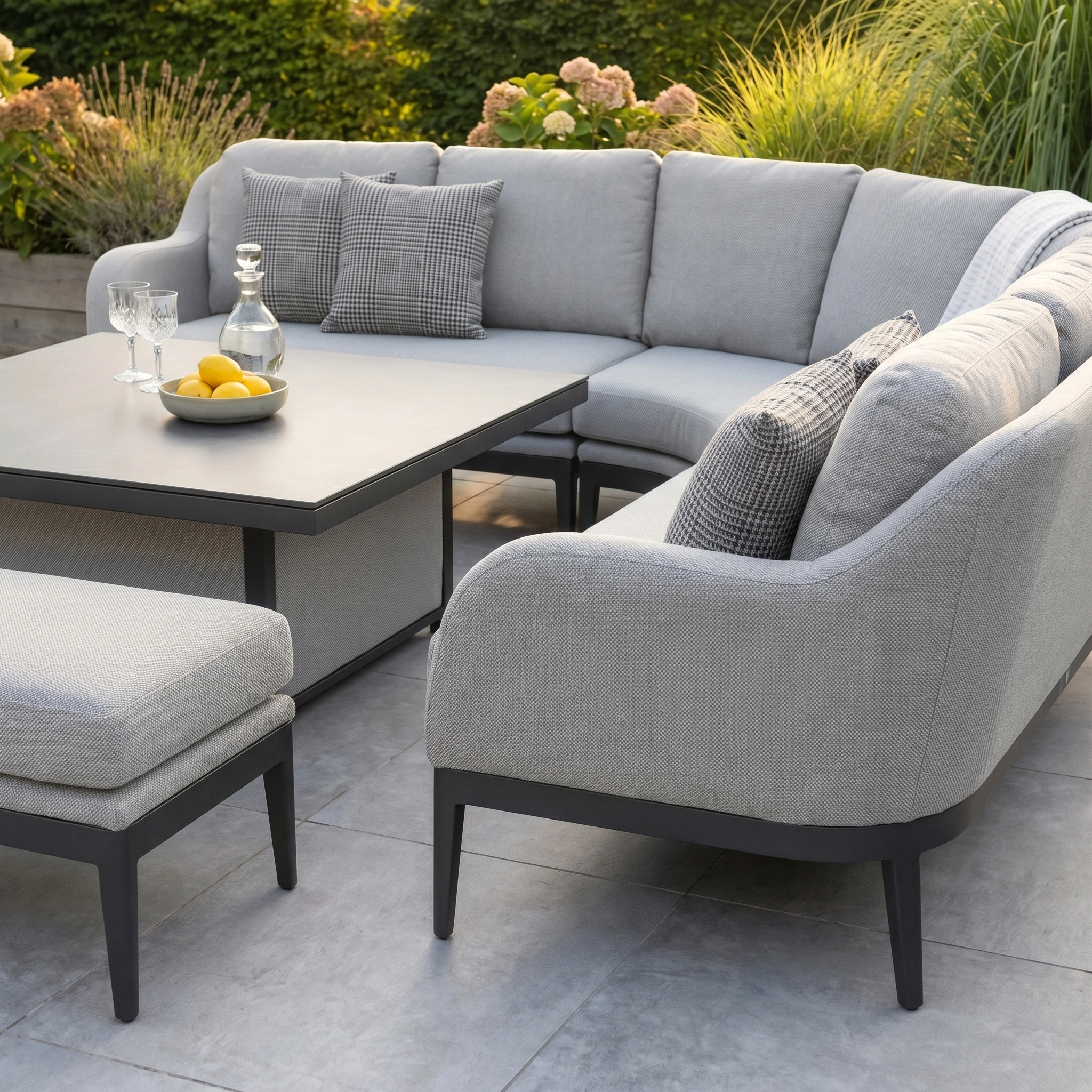 The Luna Deluxe Outdoor Fabric Square Corner Dining Set with Rising Table in Oyster Grey is shown with lemons, set against a backdrop of lush greenery.