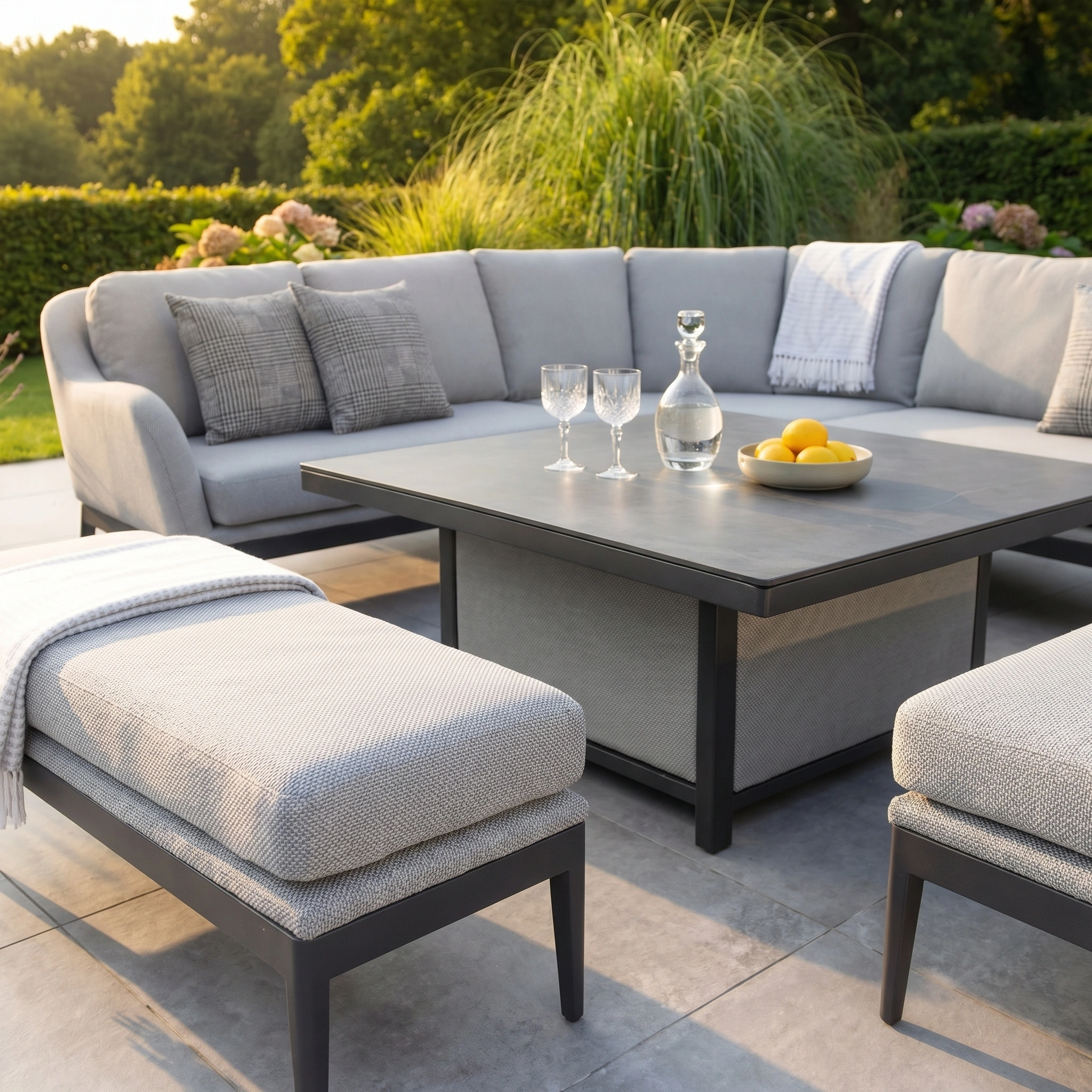 Modern outdoor patio featuring the Luna Deluxe Outdoor Fabric Square Corner Dining Set with Rising Table in Oyster Grey, complemented by glasses, a carafe, a bowl of lemons, and lush greenery.