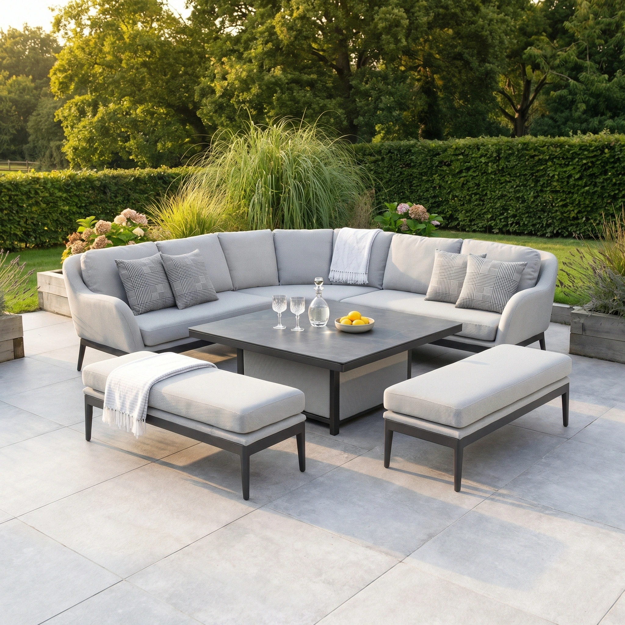 The Luna Deluxe Outdoor Fabric Square Corner Dining Set with Rising Table in Oyster Grey features cozy cushions, benches, and a versatile rising table for your patio, all set amidst lush greenery.
