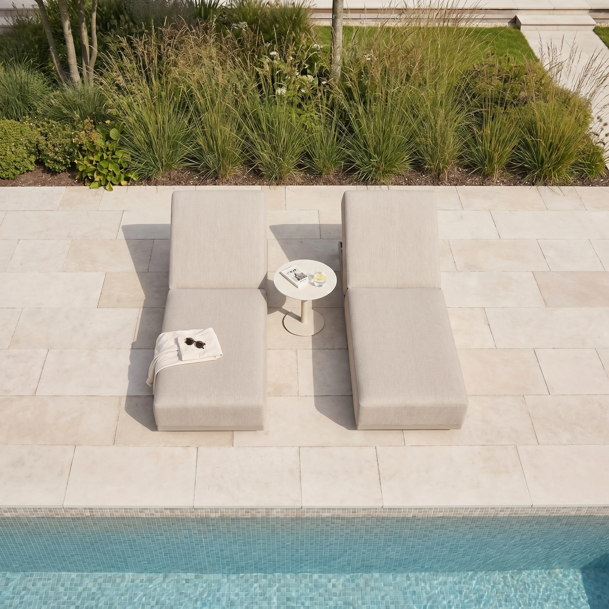 Two Luna Outdoor Fabric Deluxe Sun Loungers in Fawn sit poolside with a small round table, towel, book, and drinks between them—weatherproof outdoor furniture designed for lasting style and comfort.