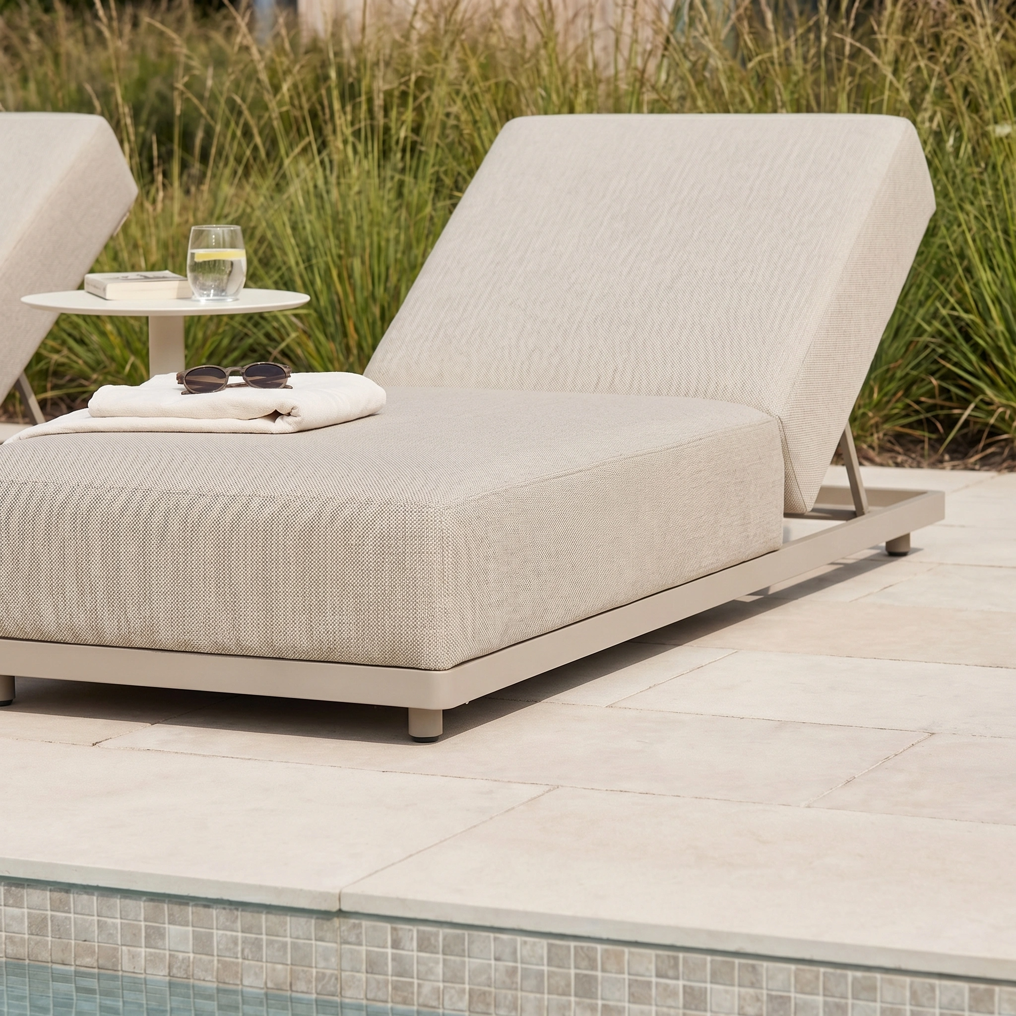The Luna Outdoor Fabric Deluxe Sun Lounger in Fawn features stain-resistant fabric and comes with a towel, sunglasses, and a glass of water on a side table—ideal, weatherproof furniture for poolside relaxation.