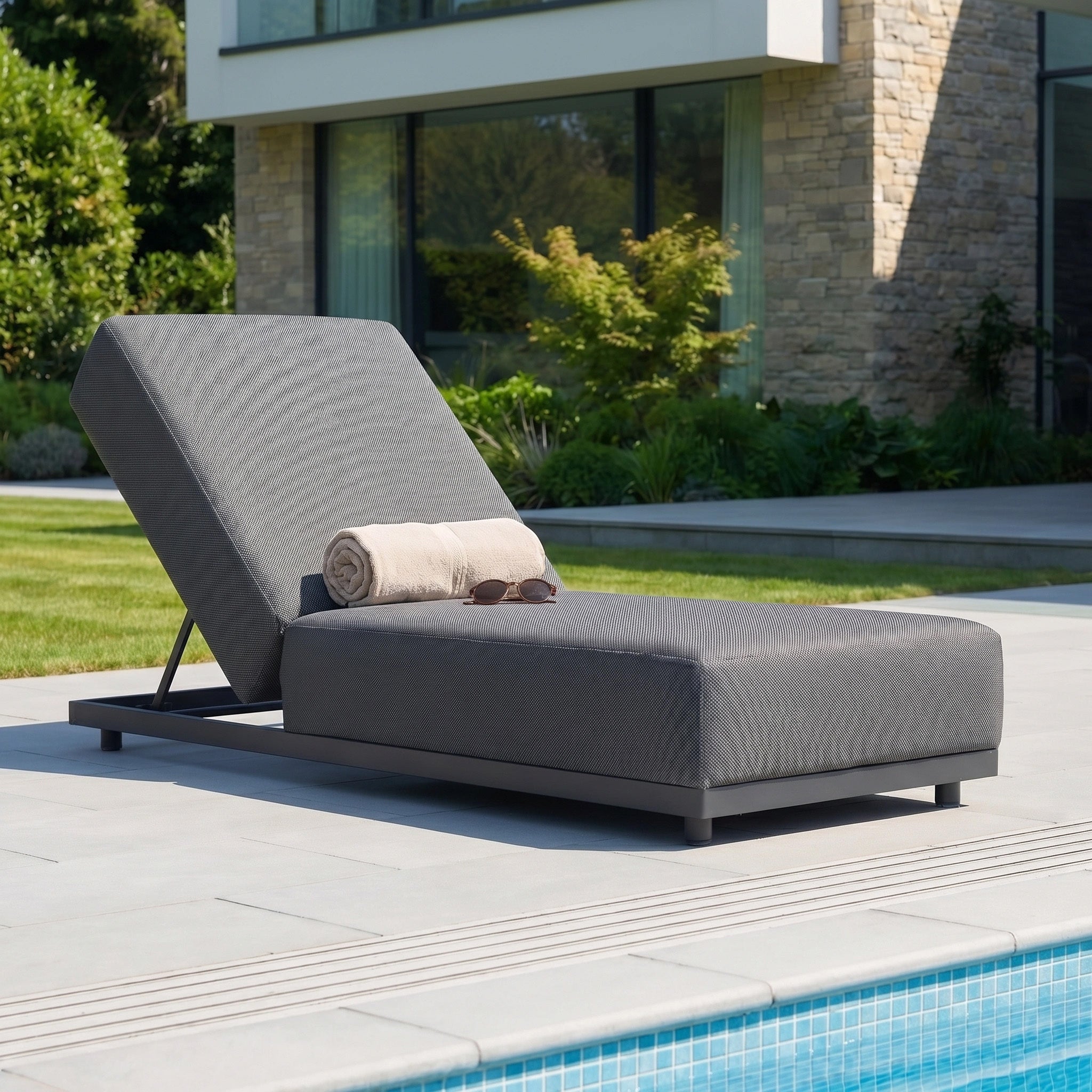 The Luna Outdoor Fabric Deluxe Sun Lounger in Grey, designed with waterproof garden furniture materials, is set poolside with a towel and sunglasses in front of a modern house.
