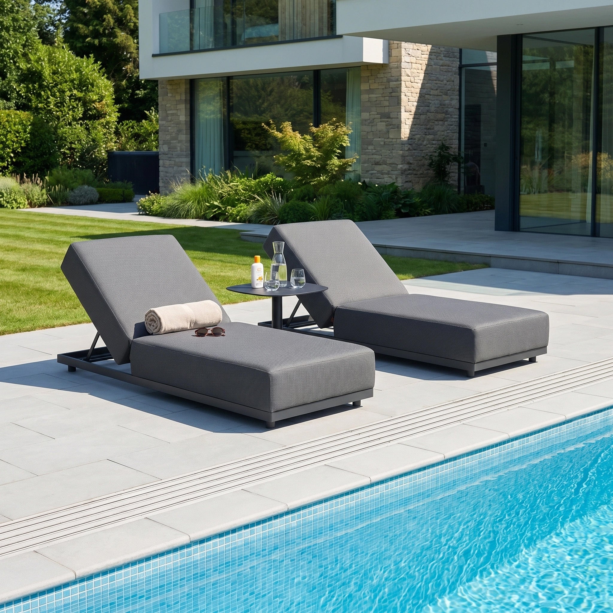 Two Luna Outdoor Fabric Deluxe Sun Loungers in Grey, made from waterproof and durable fabric, are placed with a small table beside a modern house’s pool on a sunny day.