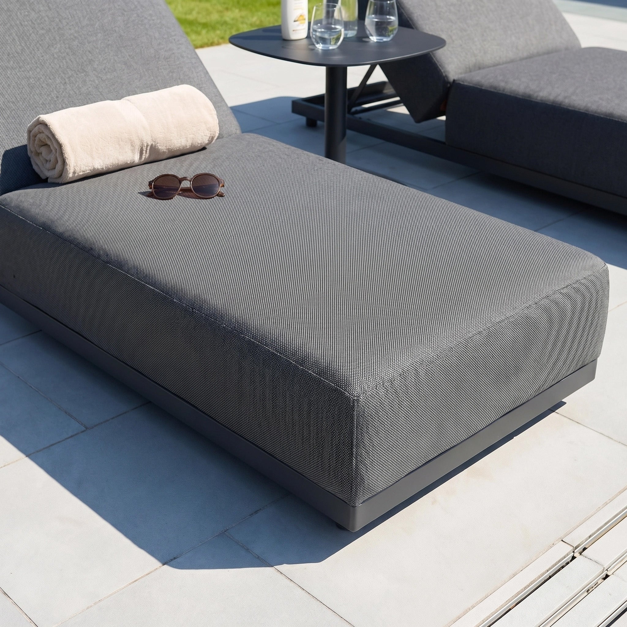 The Luna Outdoor Fabric Deluxe Sun Lounger in Grey, crafted from durable outdoor fabric, features a rolled towel and sunglasses on it and is placed beside a small black table with glassware.