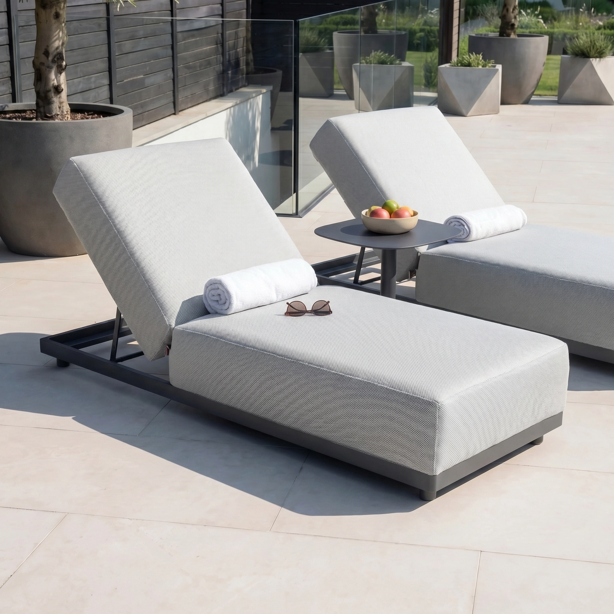 Two Luna Outdoor Fabric Deluxe Sun Loungers in Oyster Grey with towels, sunglasses, and a side table with apples sit on a sunlit patio.