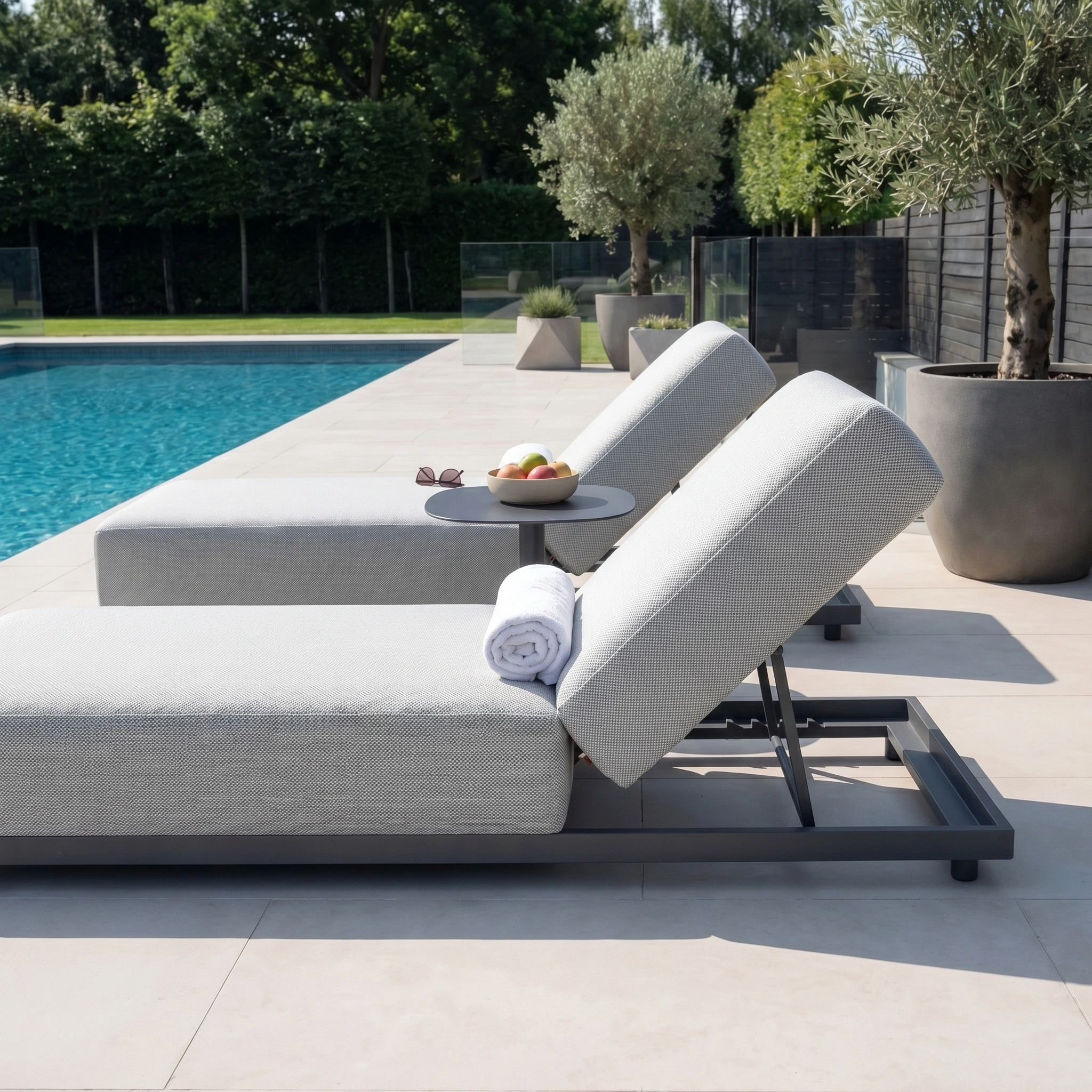 Two Luna Outdoor Fabric Deluxe Sun Loungers in Oyster Grey, paired with towels, a side table, and a bowl of fruit, create a stylish garden setup beside the pool.