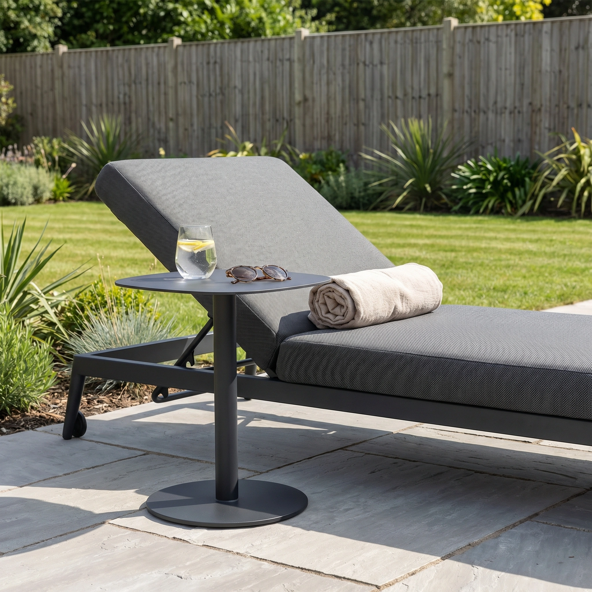 A gray outdoor lounger with a rolled towel, sunglasses, and a drink on the Luna Side Table (45cm x 45cm) in a sunny, maintenance-free garden.