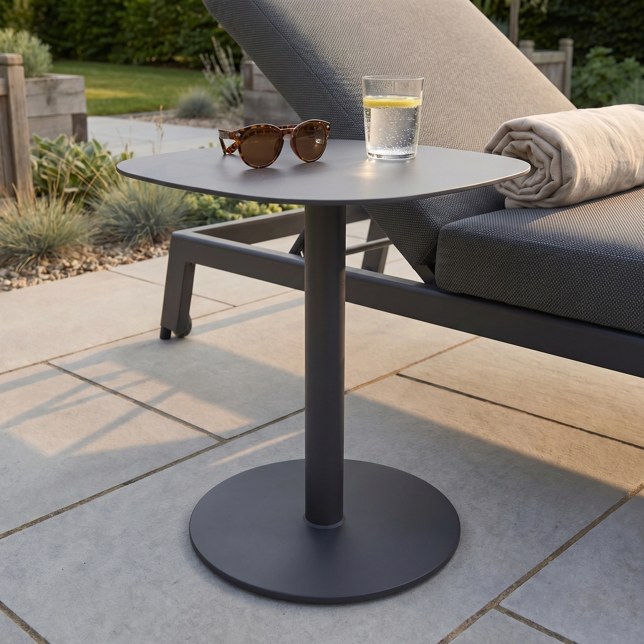 The Luna Side Table - 45cm x 45cm is a small, round, weatherproof outdoor table shown with sunglasses and a glass of water beside a lounge chair with a rolled towel.