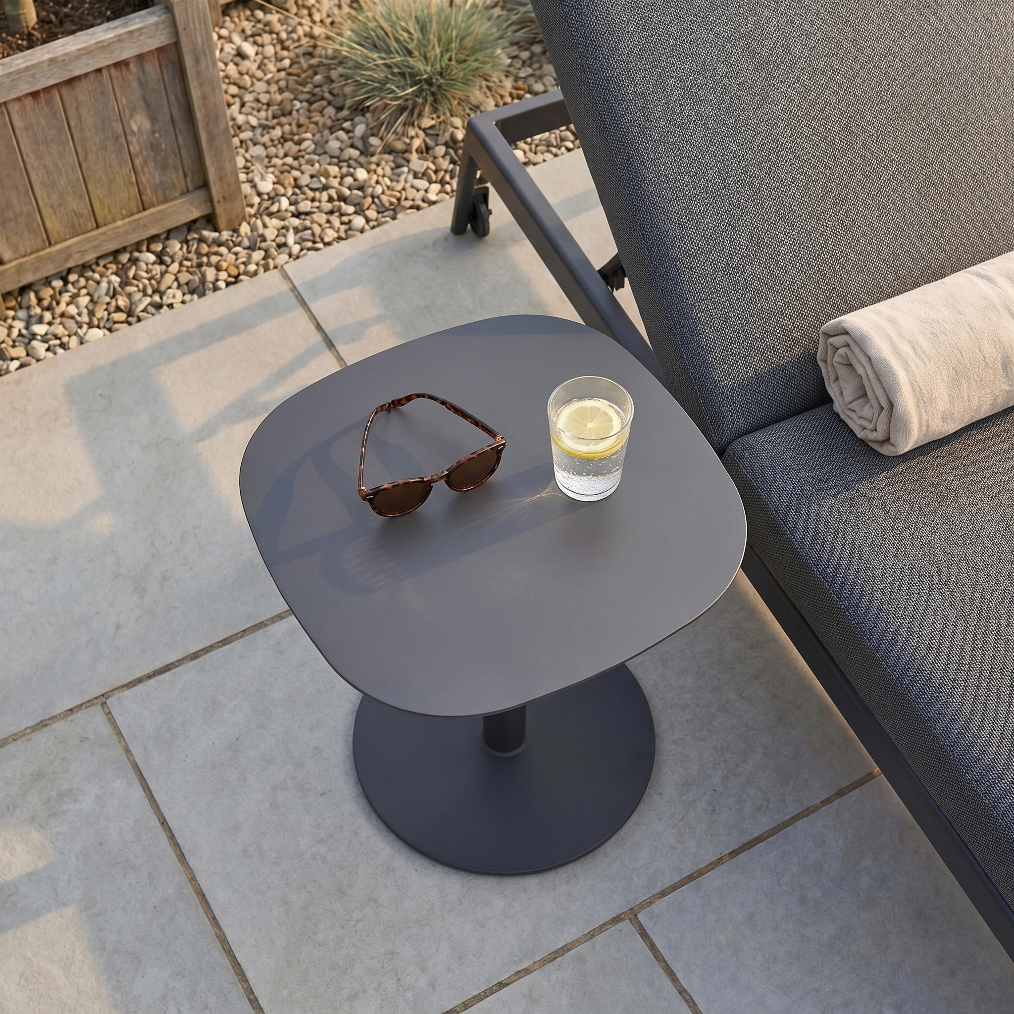 The Luna Side Table (45cm x 45cm) sits beside a gray lounge chair with a rolled towel, sunglasses, and a glass of water—ideal for maintenance-free garden furniture enthusiasts.