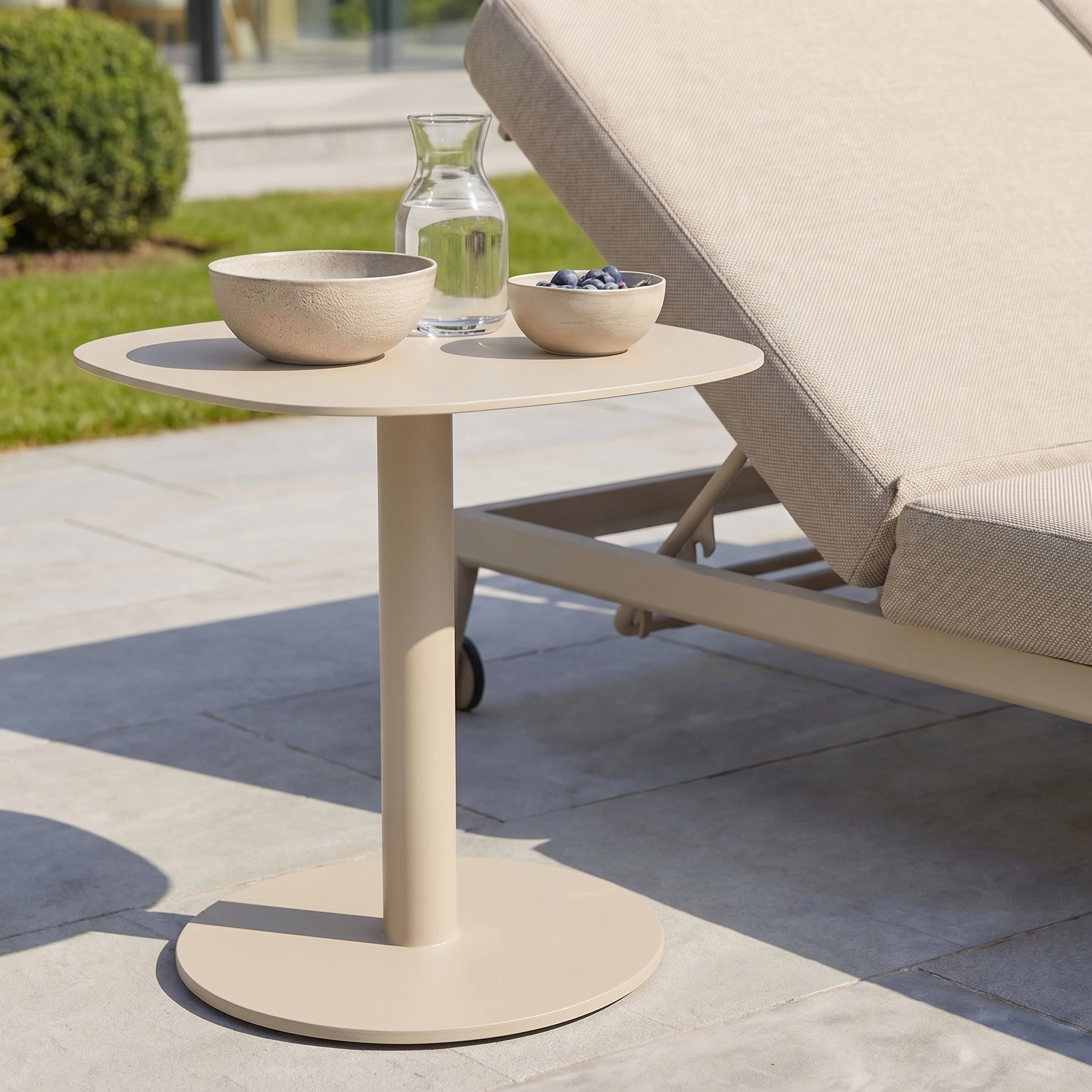 The Luna Side Table in Latte (45cm x 45cm), round and weatherproof, sits beside a lounge chair outdoors, topped with a bowl, plate of blueberries, and water carafe.