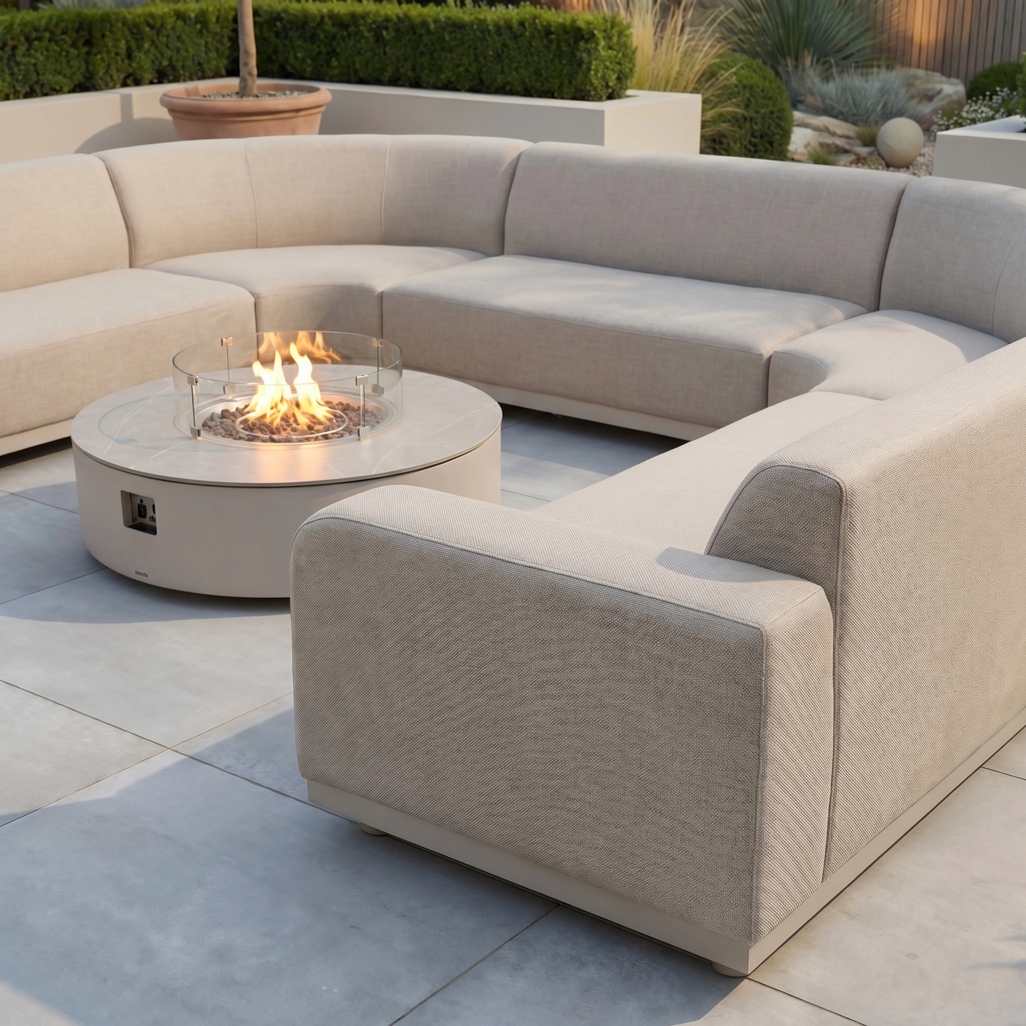 The Luna U-Shape Outdoor Fabric Curved Corner Group with Round Firepit Coffee Table in Fawn sits on a tiled patio amid lush greenery—ideal for creating stylish and inviting outdoor living arrangements.