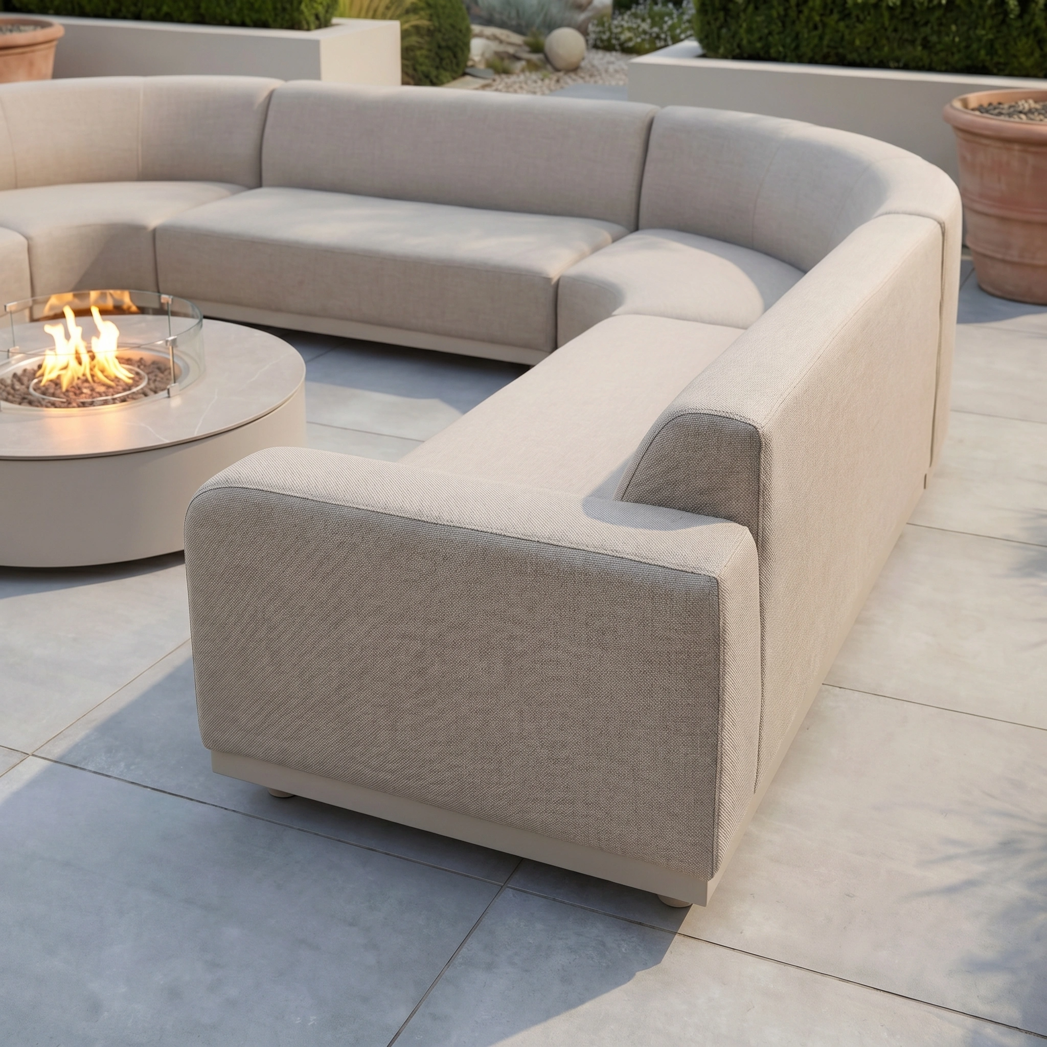The Luna U-Shape Outdoor Fabric Curved Corner Group with Round Firepit Coffee Table in Fawn is arranged on a tiled patio, with potted plants in the background.