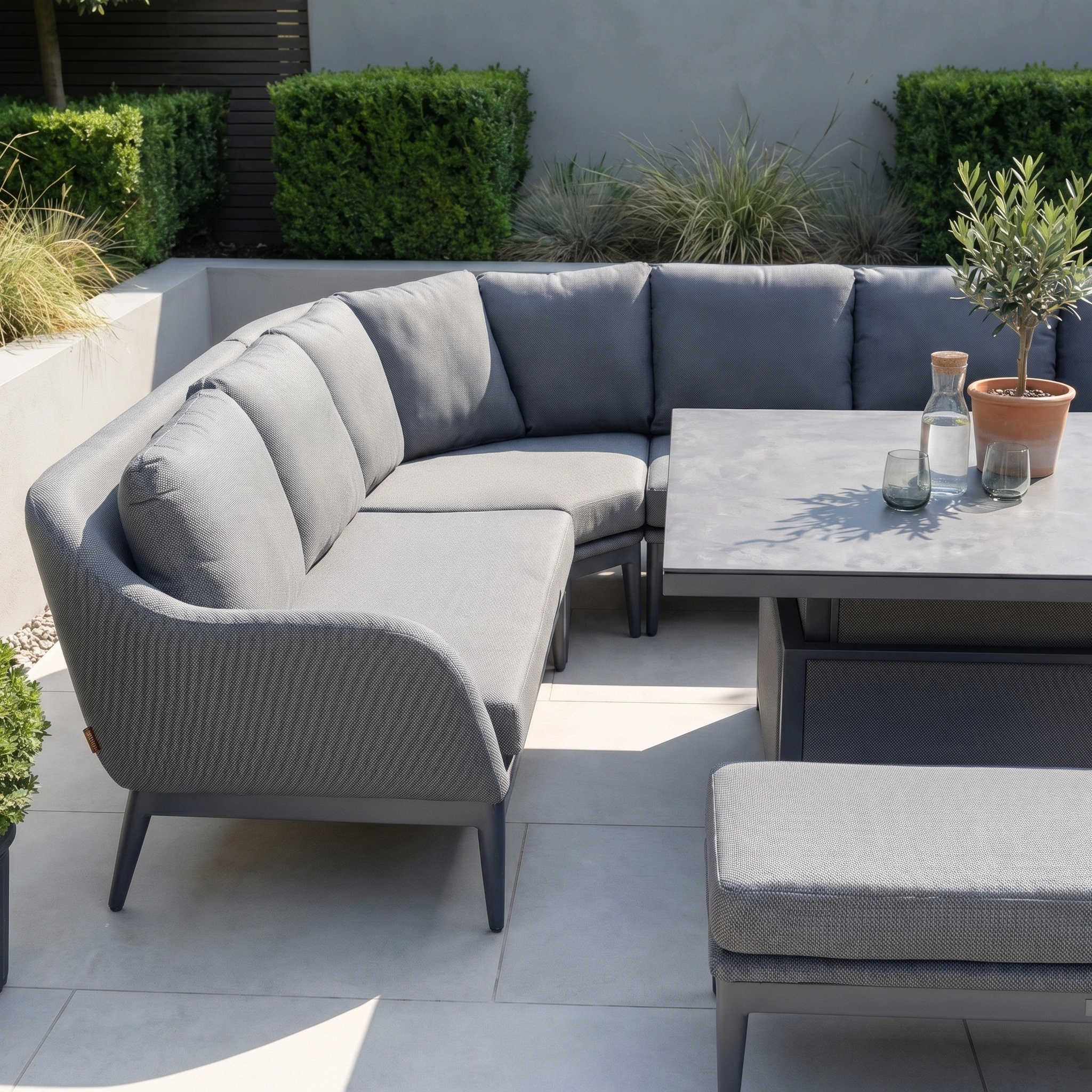 The Luna U-Shape Outdoor Fabric Corner Dining with Rising Table in Grey is arranged on a tiled patio, surrounded by green plants and shrubs.