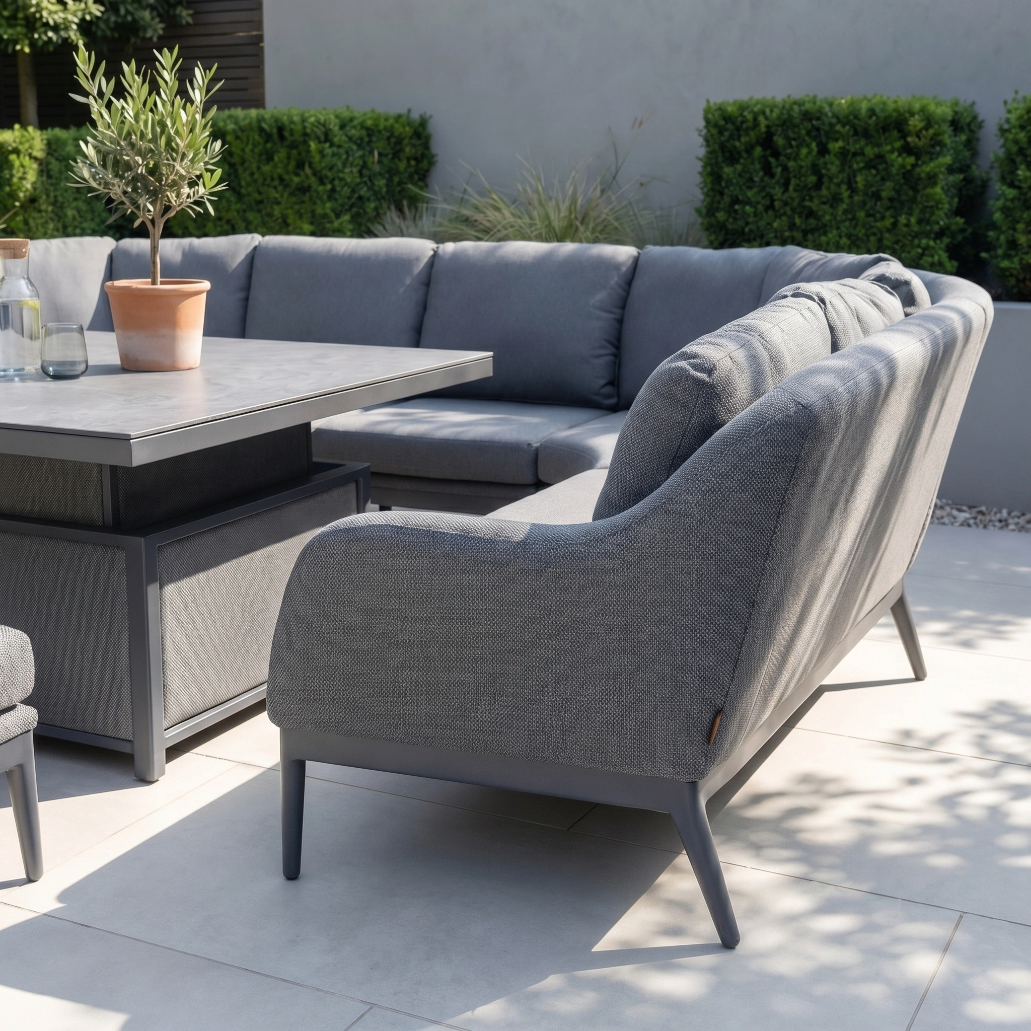 The Luna U-Shape Outdoor Fabric Corner Dining with Rising Table in Grey sits on a sunlit patio, paired with a potted plant and table, surrounded by lush greenery.