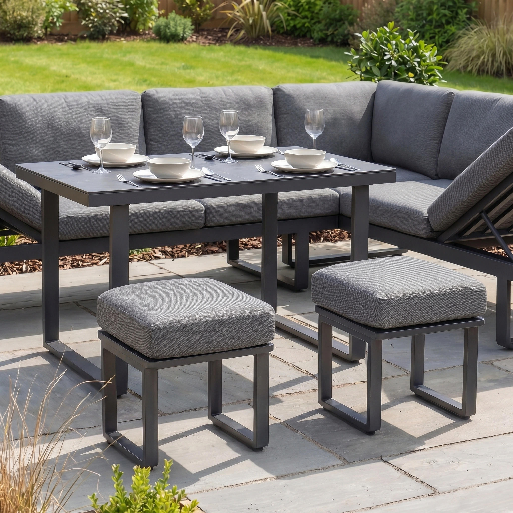 Lyra Corner Dining Set in Charcoal features gray cushions, a sturdy aluminium frame, and a table for four—ideal garden furniture with a lush green garden backdrop.