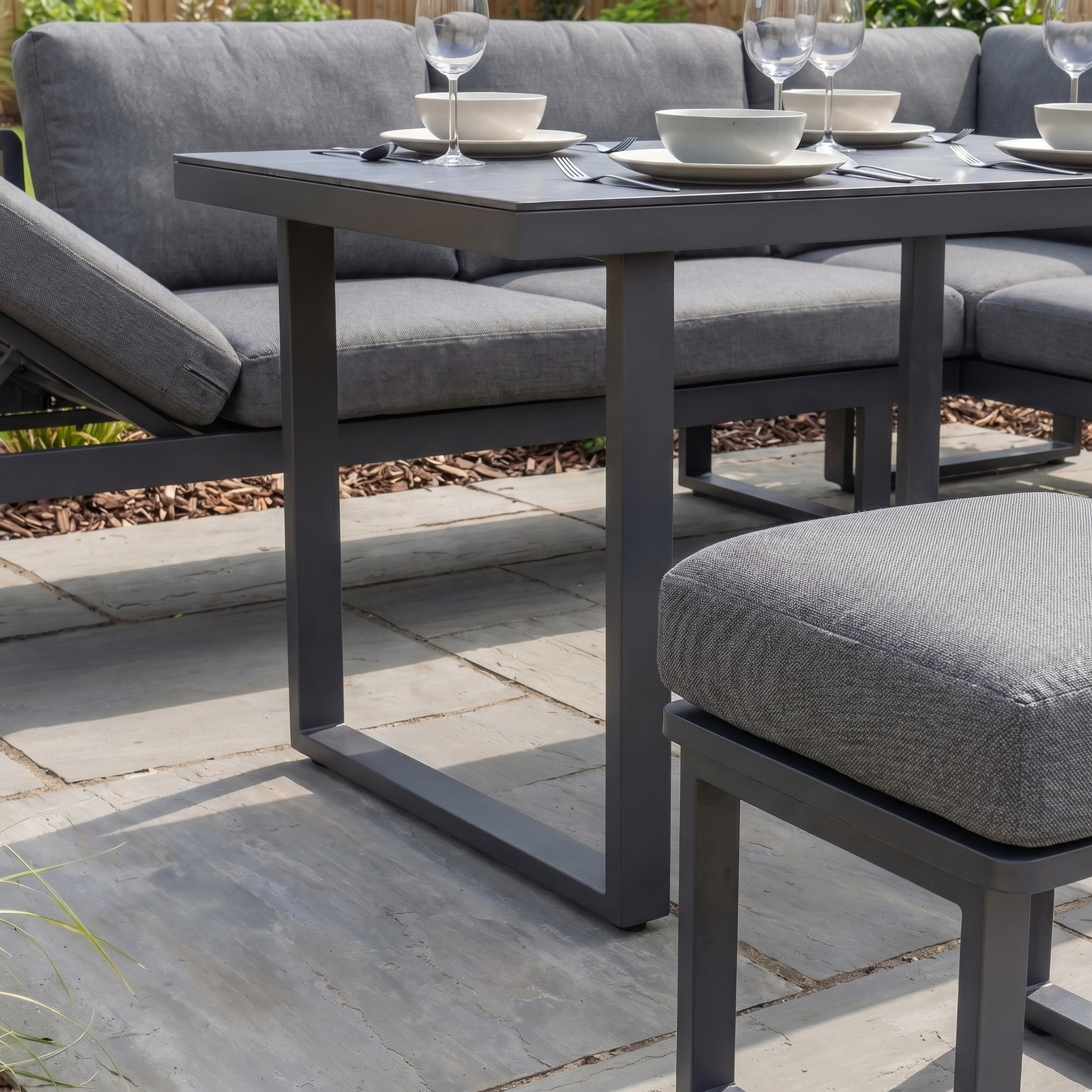The Lyra Corner Dining Set in Charcoal features gray cushioned seating and a meal-ready table on stone tiles, with a sleek aluminium frame—ideal garden furniture for relaxing outdoors.