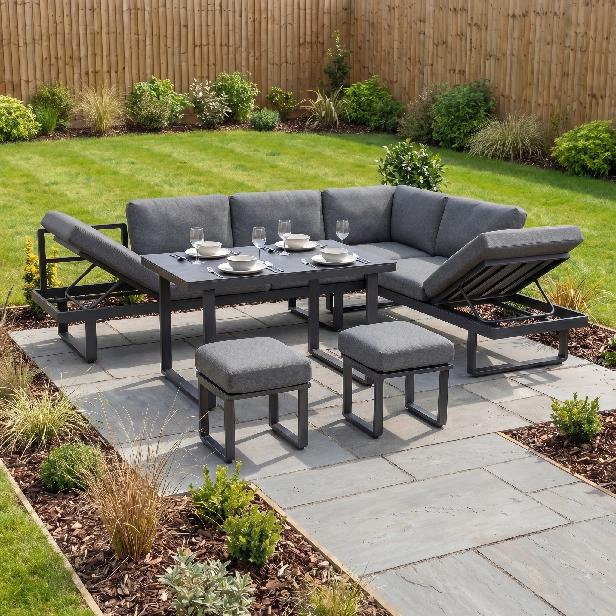 The Lyra Corner Dining Set in Charcoal, with gray cushions and a durable aluminium frame, comfortably seats four on stone paving in a beautifully landscaped garden.