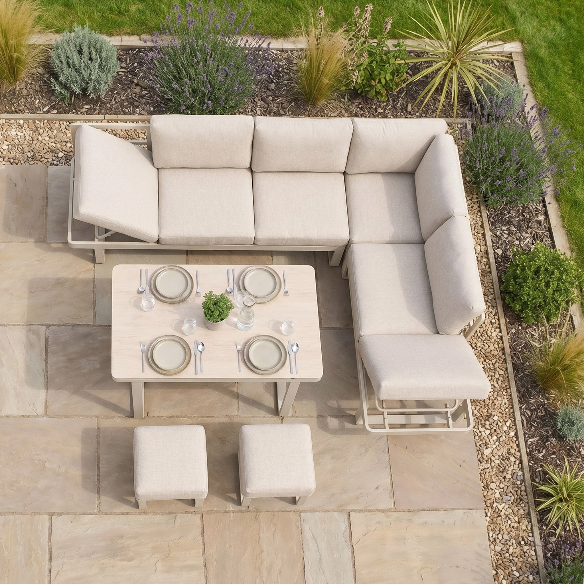 Outdoor patio with the Lyra Corner Dining Set in Latte—includes beige aluminum sofa, two stools, and a table for four—surrounded by plants and stone tiles. A perfect showcase of stylish outdoor furniture.