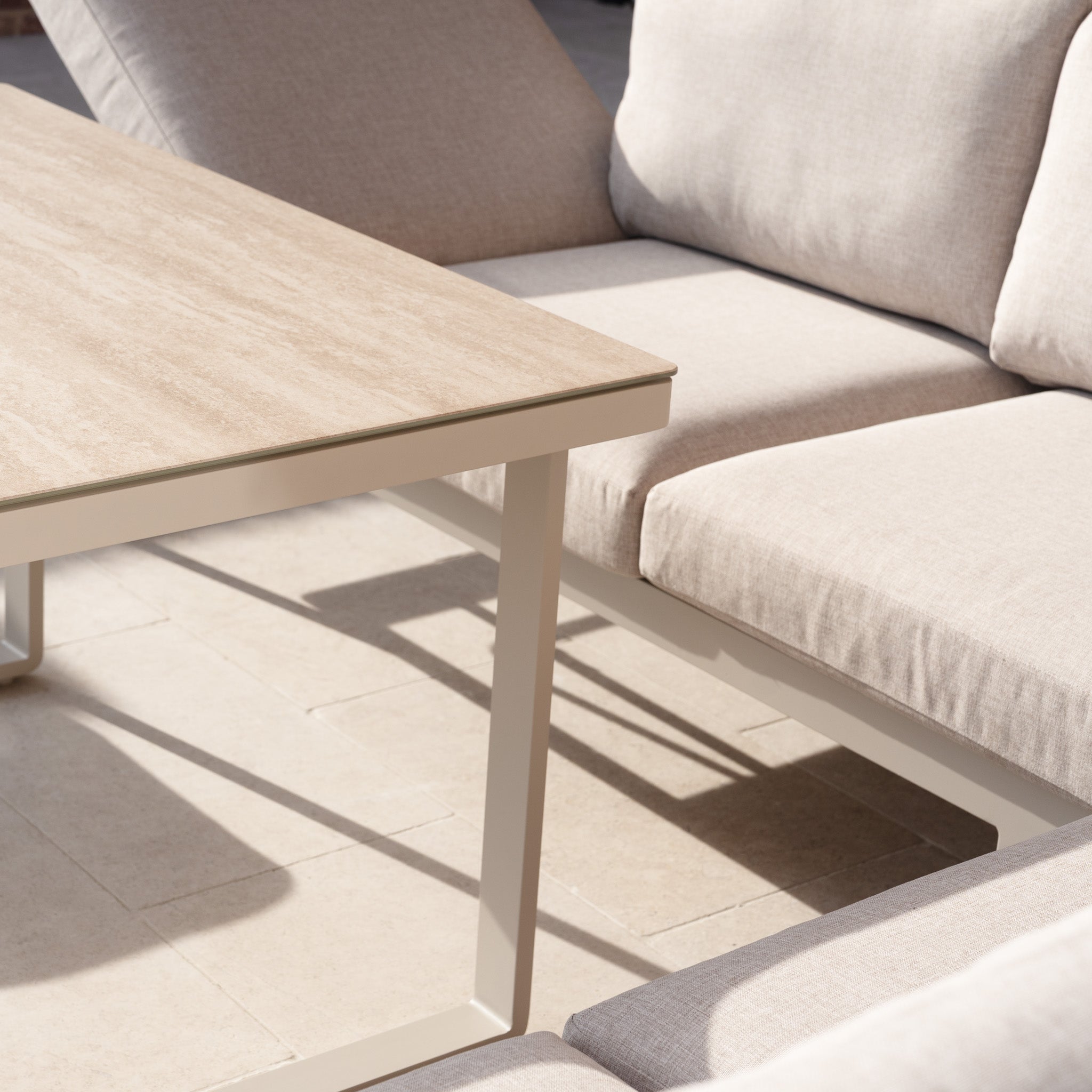 The Lyra Corner Dining Set in Latte, featuring a beige aluminium sofa and a rectangular wooden table, creates a stylish outdoor setting on a sunlit tiled patio.
